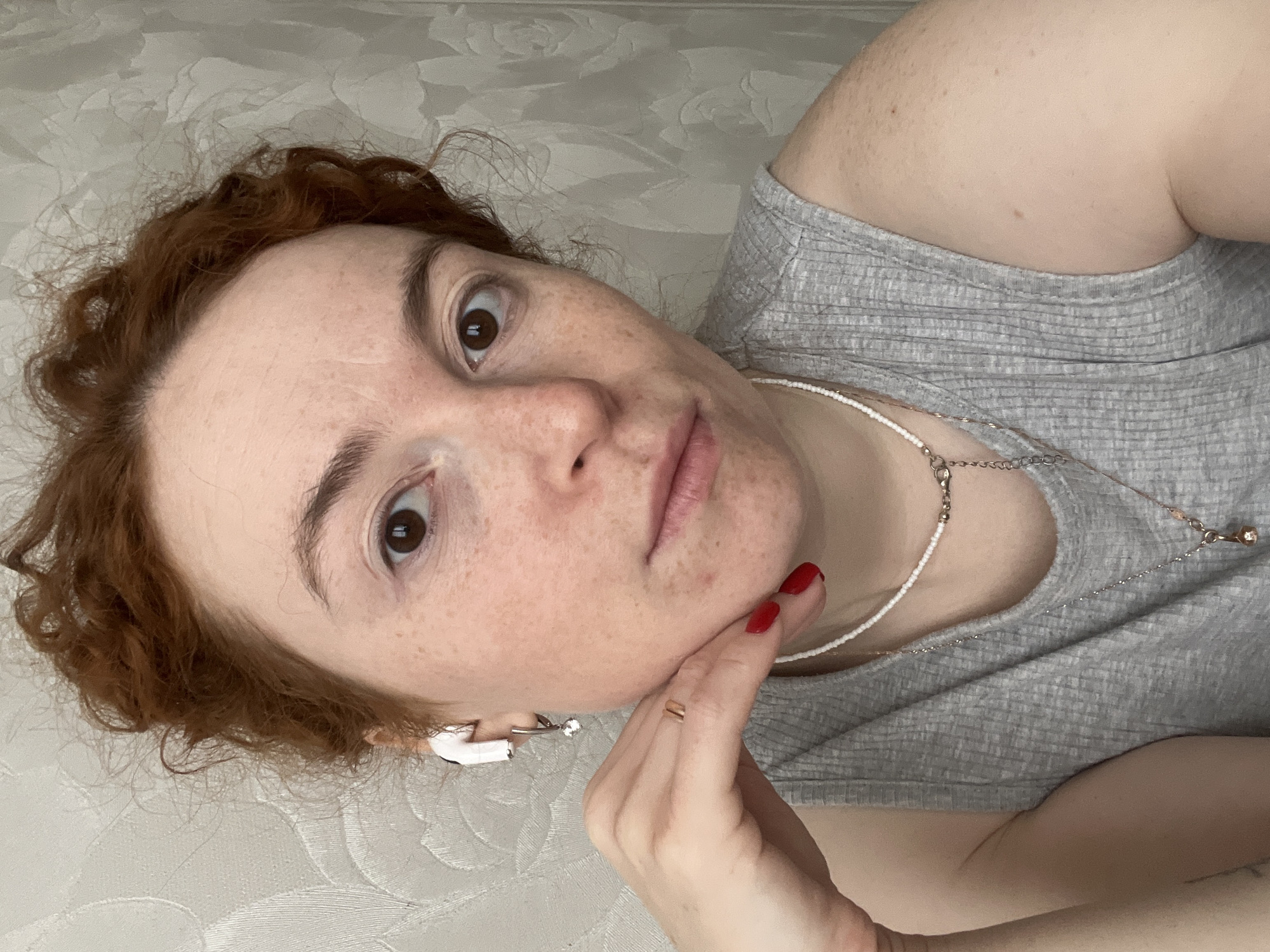 A fair-skinned person with freckles and auburn curly hair is looking at the camera. They are wearing a gray ribbed tank top and two necklaces: a short beaded white necklace and a longer, delicate silver chain necklace. The person is wearing red nail polish. They are lying on a patterned white surface, possibly a bed or couch. An earbud and a dangling earring are visible in their left ear. The person's right hand is touching their chin. There is no visible information indicating the location is Chaikovsky, Russia.