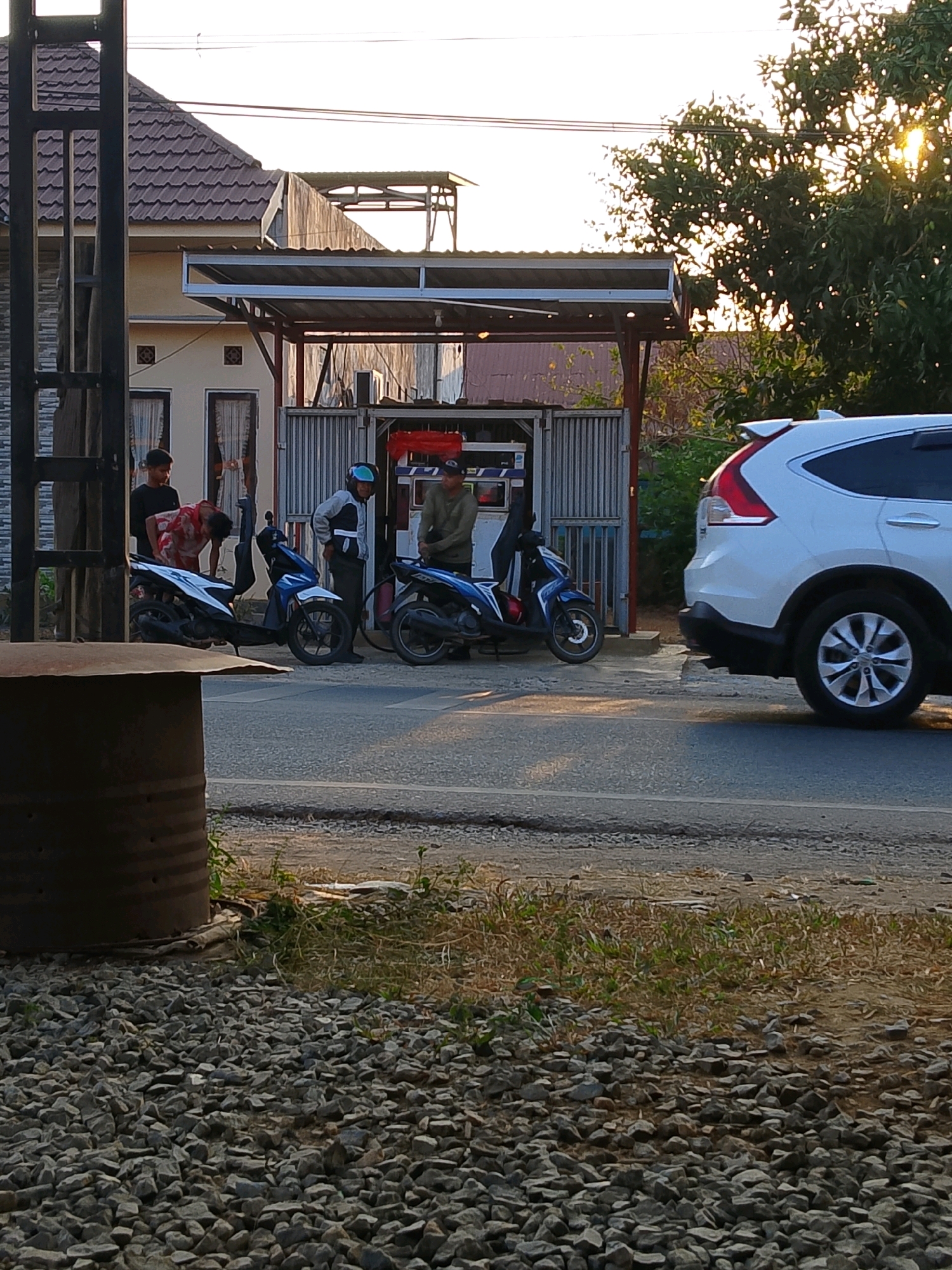The image depicts a street scene in Pelaihari, Indonesia, at what appears to be a small, open-air gas station. In the foreground, on the left, is a large, rusted metal barrel situated on a bed of gravel. A paved road runs through the middle of the image, with a white SUV visible on the right side, parked partially on the shoulder.
The gas station itself is a modest structure with a corrugated metal roof supported by red posts. A white and blue structure, likely a fuel pump, is visible within the station. Several people are gathered around motorcycles in front of the station.
On the left side of the gas station, a person in a red and white shirt is bending over a blue and white motorcycle, seemingly engaged in refueling. Another person in a black shirt stands behind them. To the right of this, two men are interacting near another motorcycle. One man, wearing a helmet and a grey and black jacket, appears to be standing by a blue motorcycle. Another man in a light green long-sleeved shirt and a black cap is near the fuel pump, holding a nozzle, presumably refueling the motorcycle he is near.
The background shows a building with a tiled roof, an outdoor area with some greenery and trees, and power lines overhead. The lighting suggests it is either early morning or late afternoon, with the sun casting a warm glow. The overall atmosphere is that of a casual, everyday transaction at a local service station.