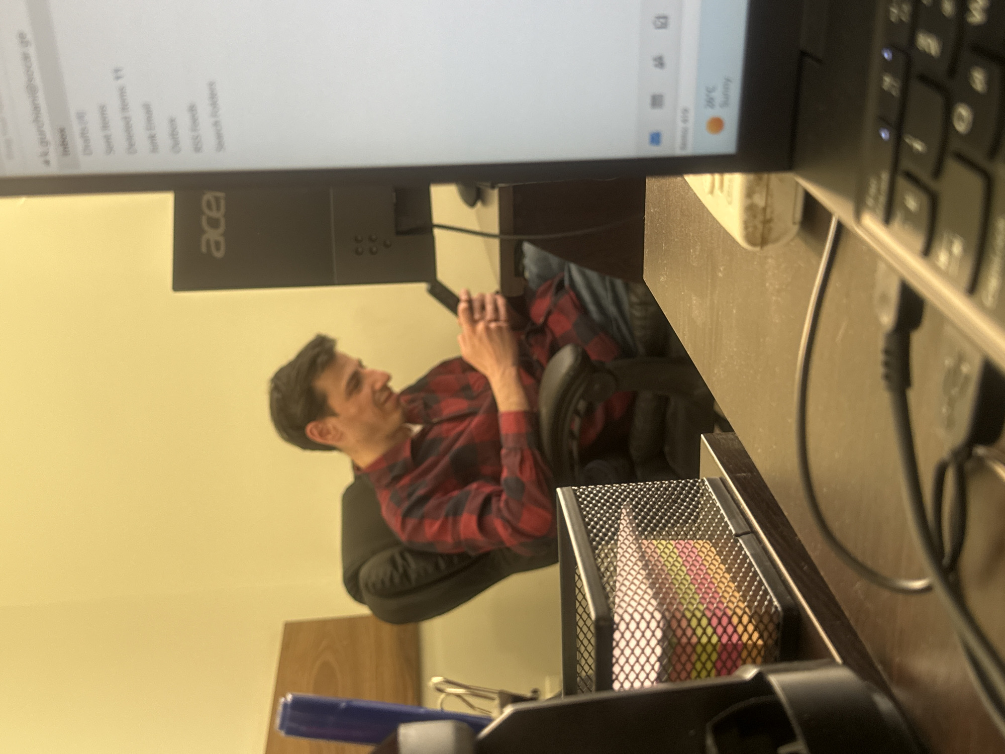 The image depicts a casual moment in an office setting in Tbilisi, Georgia. A man with dark hair, wearing a red and black plaid shirt, is seated in a black office chair, leaning back with a relaxed and smiling expression. His gaze is directed upwards and to the side, and his hands are positioned near his stomach, possibly holding a mobile device or resting.
In the foreground, a laptop is partially visible, showing its screen and a portion of its keyboard. The screen displays an email application interface with an email address "k.gurchiani@socar.ge" and folders such as "Inbox," "Drafts (4)," "Sent Items," "Deleted Items 11," "Junk Email," "Outbox," "RSS Feeds," and "Search Folders." The screen also indicates "20°C Sunny" as the current weather. Behind the laptop, a black Acer brand monitor is visible.
The desk surface is dark and appears slightly dusty. Notable objects on the desk include a white power adapter, various black cables, and a black mesh desk organizer filled with colorful sticky notes in shades of pink, yellow, and green. A blue pen or marker is also seen in the lower foreground. The background features a plain light yellow wall. The scene suggests a relaxed atmosphere in a personal workspace, with the man taking a break or engaged in light activity.
