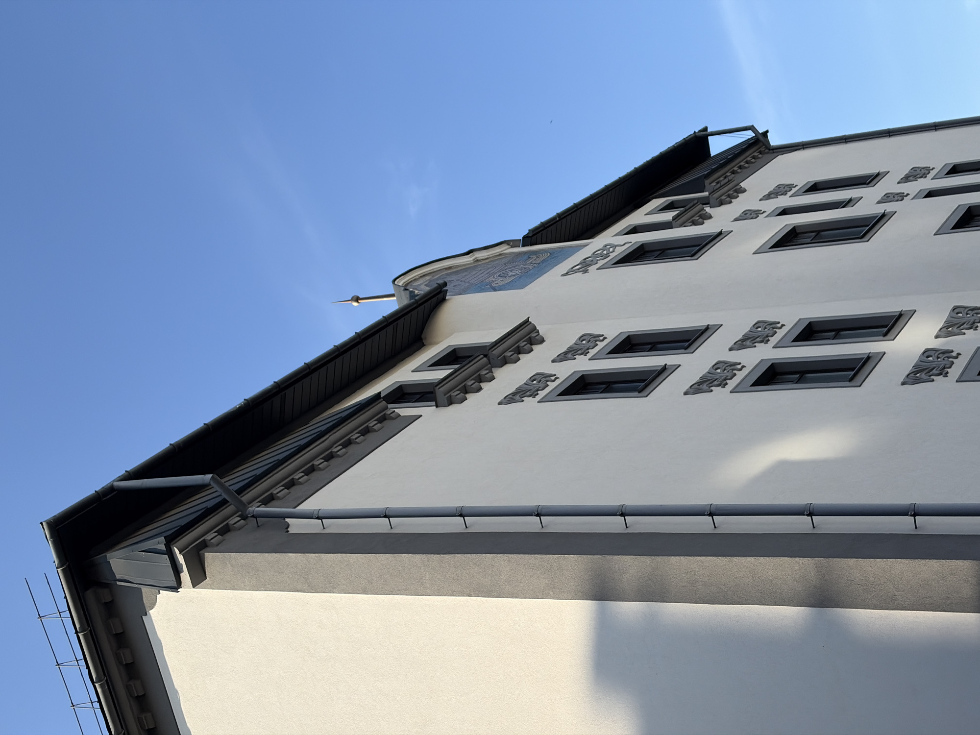 An exterior, low-angle, Dutch tilt shot captures the side of a white and grey building against a clear blue sky. The building features numerous dark-framed windows and decorative grey elements above some of them. A dark grey downspout runs vertically along the side of the building, attached with black brackets. The roof is dark grey with black trim. Shadows from the building and a railing are cast on the left side of the facade. In the distance, a small airplane is visible in the sky. The architectural style of the building, with its ornate window decorations and mansard-style roof, is characteristic of buildings found in the historic center of Minsk, Belarus, particularly around the Upper Town district.