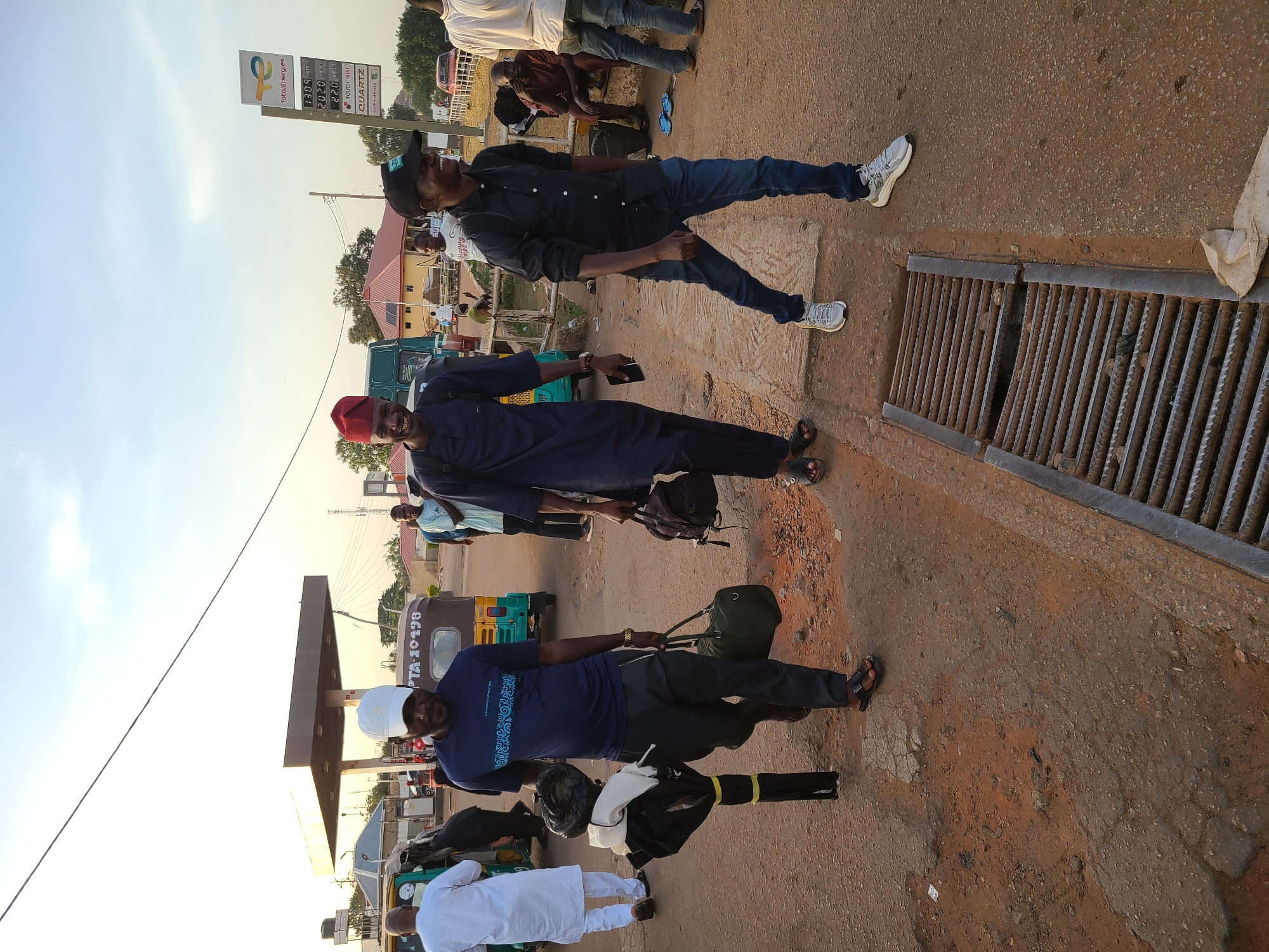 This media file captures a vibrant street scene in Jos, Nigeria, during the daytime under a clear sky. The setting appears to be a busy roadside adjacent to a TotalEnergies filling station, evidenced by the sign visible in the background.
Several men are the primary subjects. In the foreground, a man wearing a white cap, a dark blue t-shirt with a pattern that includes "LUMUMBA UNIVERSITY ATHLETICS", black trousers, and sandals holds a green duffel bag and another black item. Beside him, another man in a red cap, a dark blue tunic, and sandals smiles while holding a mobile phone. Further back, a man in a black button-down shirt, blue jeans, white sneakers, and a black cap is also seen smiling. Other individuals are visible in the background, some standing, some sitting, and one in white traditional attire.
The activity suggests a casual gathering, possibly people waiting for transport, conversing, or pausing at the gas station. Vehicles present include a distinctive green and yellow tricycle (commonly known as 'keke napep') with the license plate "PTA-10198" clearly visible, along with motorcycles and other cars.
Notable details include the bright, clear weather, indicating a sunny day. A prominent TotalEnergies sign displays fuel prices: PETROL at 130.9, DIESEL at 202.0, and KERO at 22.0. The sign also lists "TOUCH POINT" and "QUARTZ". A metal drainage grate runs along the roadside. The general ambiance points to a bustling public area or transport hub within an urban or semi-urban environment.
