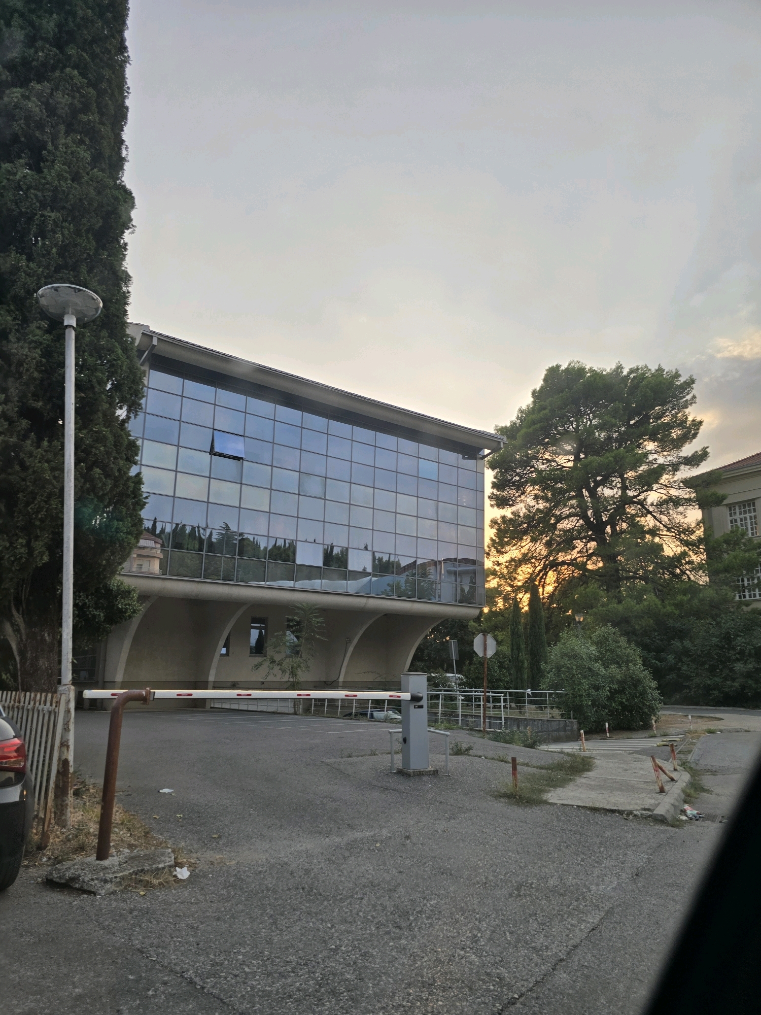The image captures a modern building with a glass facade, partially obscured by large trees. In the foreground, a barrier gate is visible, suggesting an entrance or checkpoint. The sky is overcast with a hint of sunset glow, and reflections of trees and buildings can be seen on the glass of the structure. The scene appears to be an outdoor setting, possibly a campus or office park, given the architecture and landscaping. The location is identified as the University of Montenegro's Faculty of Law building in Podgorica, Montenegro. The image shows the building's distinctive curved understructure and the extensive use of glass panels on its upper floors. There are no people visible in the frame.