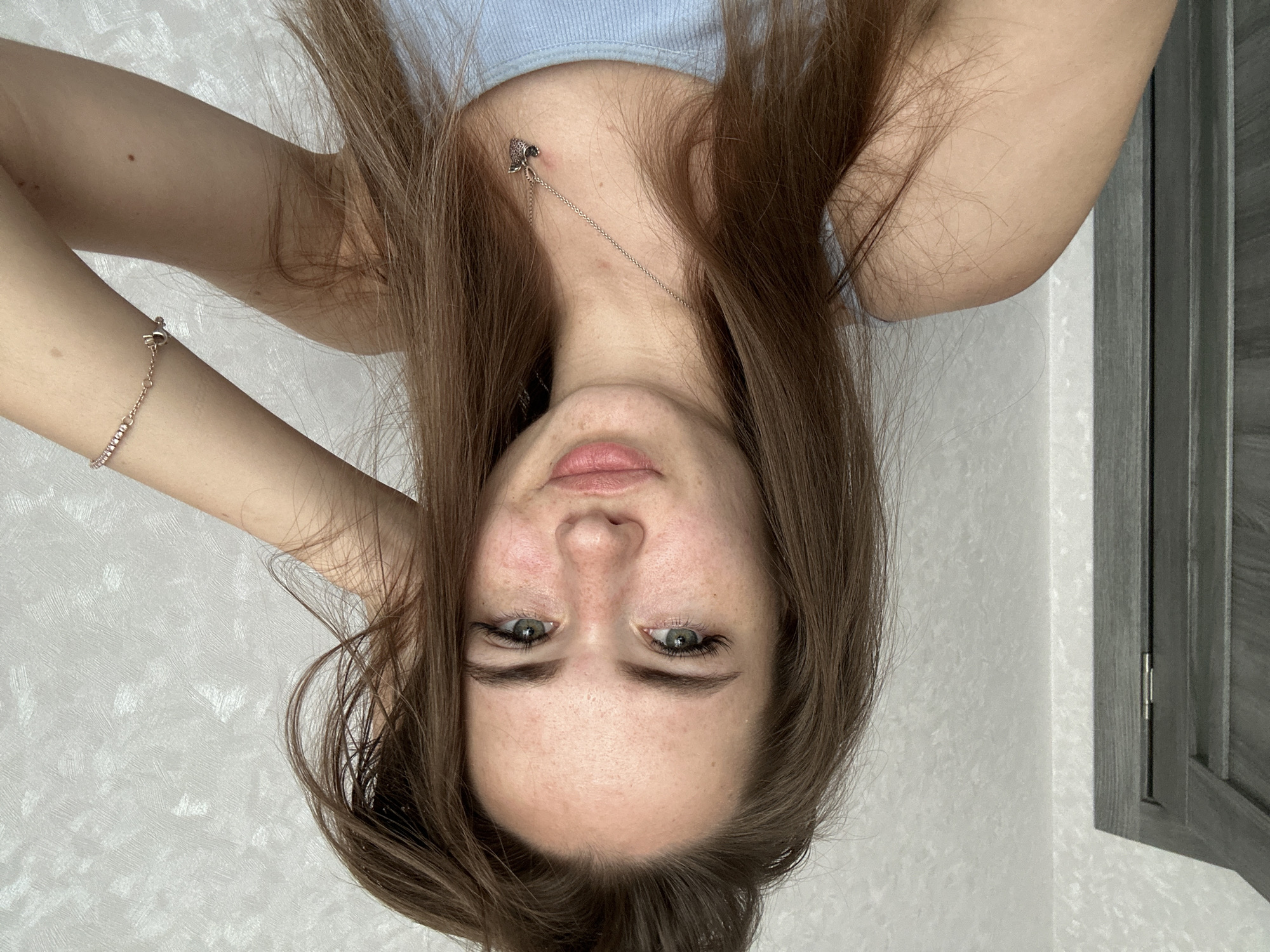A young woman with light brown hair and fair skin is taking a selfie indoors. She has green eyes and light freckles on her face. She is wearing a light blue tank top and a silver necklace with a heart-shaped pendant. Her left hand is resting on her head, and her right arm is raised, with a bracelet on her wrist. The background consists of a white textured wall and a portion of a gray door. The image is well-lit, with natural light appearing to come from the right.