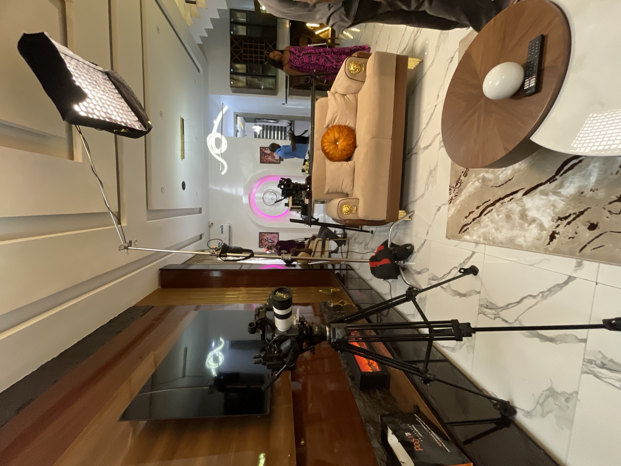 This is a medium shot of a home interior that appears to be set up for a video shoot. In the foreground, a professional video camera is mounted on a tripod. To the right of the camera, a light fixture is suspended from the ceiling. The room is furnished with a light-colored sofa, a coffee table with a decorative orb and a remote control, and a large wall-mounted television. In the background, several people are visible, including a woman in a patterned dress standing near a bar area, and two men who appear to be operating the camera equipment. The overall impression is of a modern, well-lit living space being utilized as a production set.