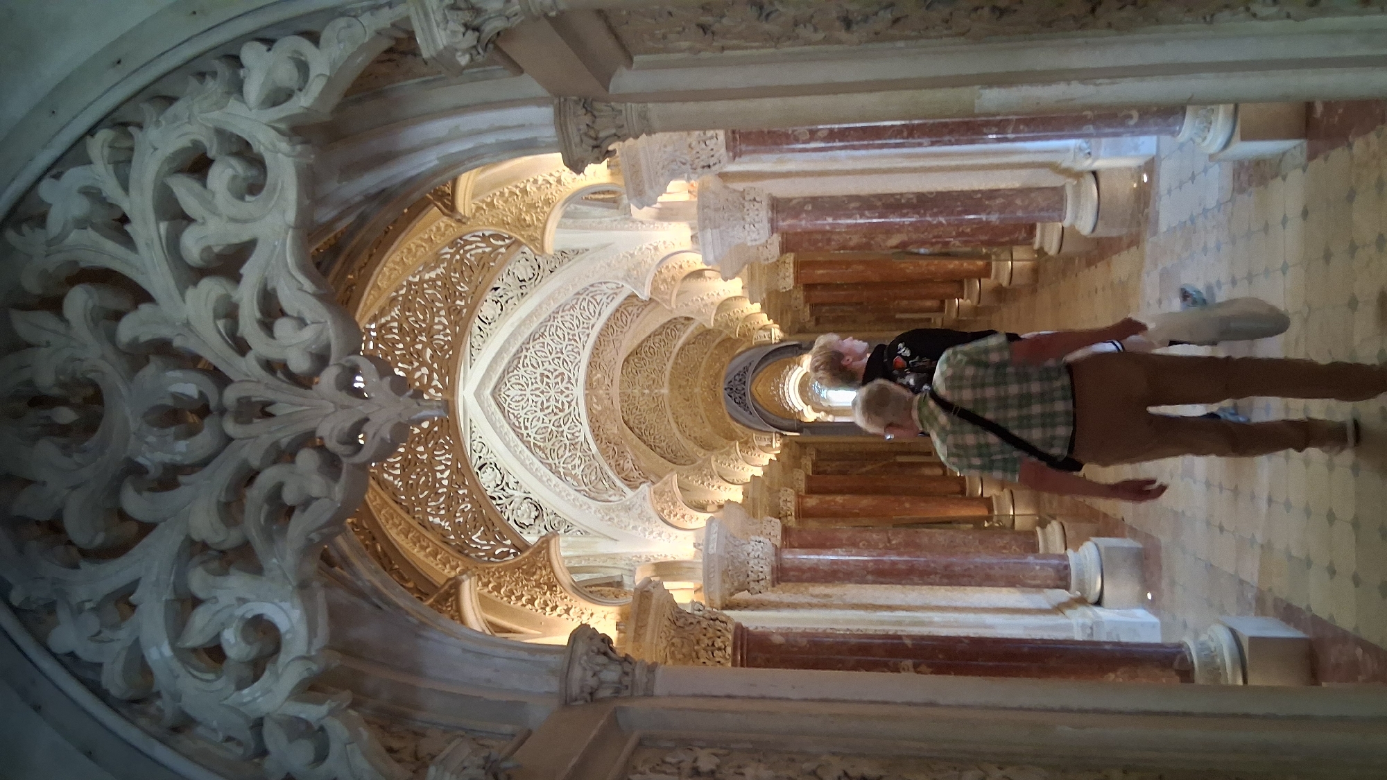Two individuals walk down a richly decorated hallway in a historical building. The hallway features a series of arches with intricate carvings and detailed soffits, illuminated by soft lighting. Marble columns, some with red and white veining, line the corridor. The floor is tiled in a checkerboard pattern. The person in the foreground is an older male wearing a green and white plaid shirt, khaki pants, and a crossbody bag. He is carrying a white shopping bag. Behind him, a younger individual with blonde hair, wearing a dark sweatshirt, walks ahead. The architecture suggests a location like Pena Palace in Sintra, Portugal.