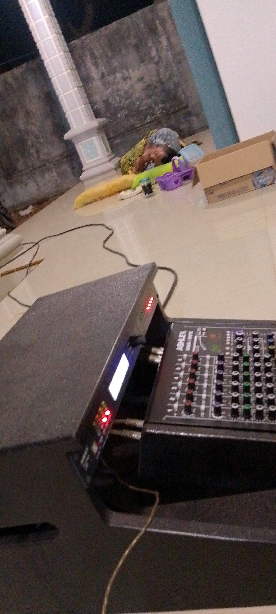 The image depicts an indoor scene with a tiled floor and a textured wall in the background. In the foreground, a large black speaker unit is visible, with a smaller black audio mixer placed on top of it. The mixer has several knobs and buttons, along with a digital display that is illuminated. Red indicator lights are also visible on the mixer and a connected black electronic device. A thin wire snakes across the floor and onto the speaker unit. In the background, a decorative white pillar stands near the wall, and several objects, including a yellow cylindrical item, a green fabric, and a purple plastic toy, are scattered on the floor. A cardboard box is also present in the background. The lighting suggests an indoor setting, possibly a room in Batubintang, Indonesia, where this equipment is set up.