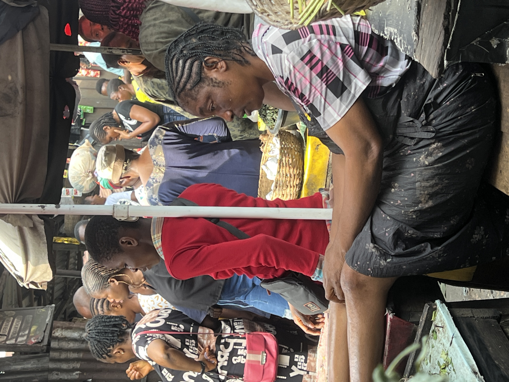 The image shows a busy market scene in Onitsha, Nigeria. In the foreground, a woman with braided hair, wearing a pink and white checkered shirt and a dark floral skirt, sits on a wooden surface. Her legs are crossed, and her arms are resting on her lap. To her left, a woman in a red long-sleeved shirt is visible, with a black bag slung across her body. Behind them, a crowd of people are milling about, some with bags and baskets. The market appears to be covered by awnings, and various goods are displayed, including produce in woven baskets. The overall atmosphere is one of vibrant commerce.