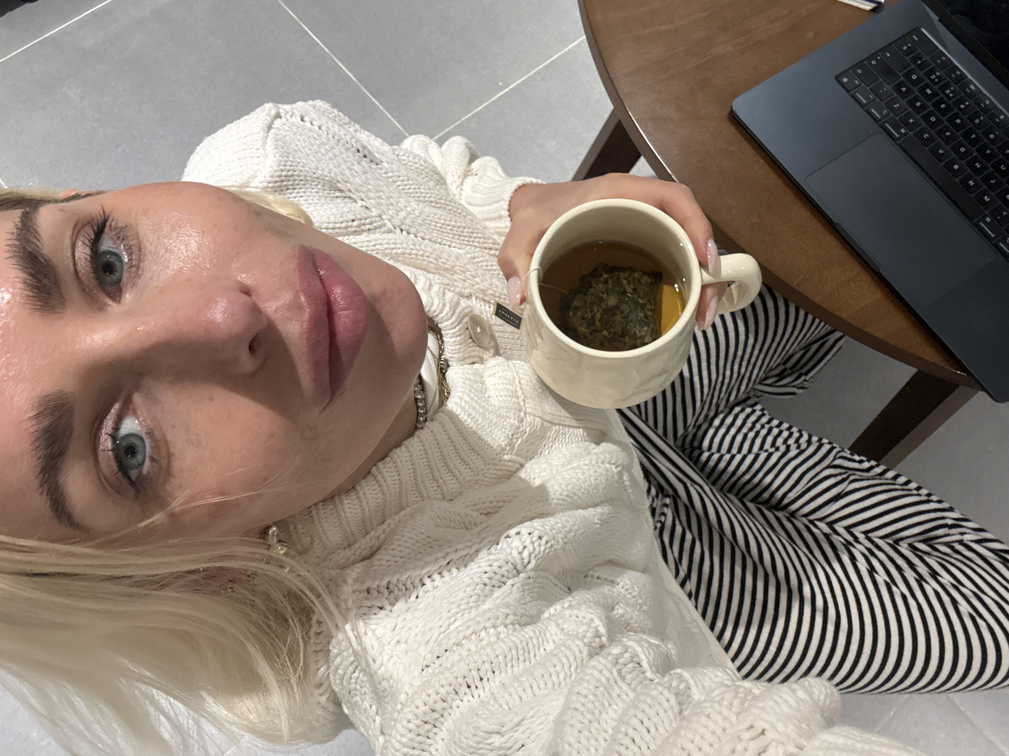 A person with blond hair and blue eyes holds a mug of tea while sitting near a laptop on a small wooden table. The person is wearing a white knitted sweater and striped pants. The location is unspecified, and there is no evidence to suggest this image was taken in Dubai, United Arab Emirates.