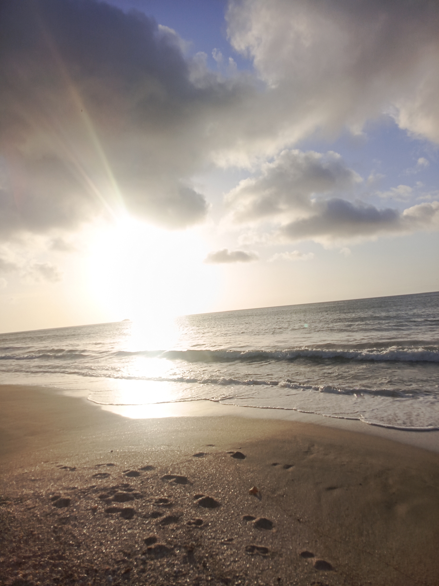 A low-angle, wide shot captures a beach scene at sunset in Pedro González, Venezuela. The sun, a bright white orb, is partially obscured by dramatic, dark grey clouds that cast a warm glow across the sky and sea. Beams of sunlight radiate from the sun, creating a lens flare effect that washes out parts of the image. The ocean stretches to the horizon, with gentle waves breaking on the shore, their foam illuminated by the setting sun. The sandy beach in the foreground is wet and reflective, showing scattered footprints. The environment is tranquil and natural, with no people or man-made structures visible.