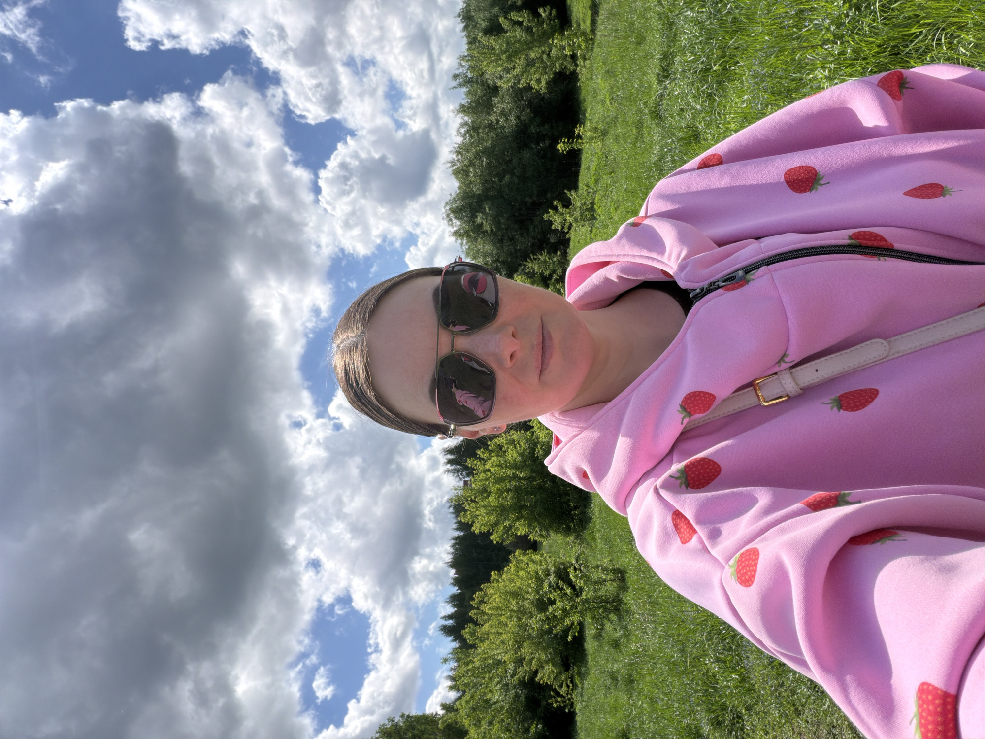 A medium shot captures a fair-skinned woman with dark hair and sunglasses, wearing a pink strawberry-patterned hoodie. She is outdoors in what appears to be a grassy field on a partially cloudy day. Trees are visible in the background behind her. The location is not specified in the city of Unknown, Unknown.