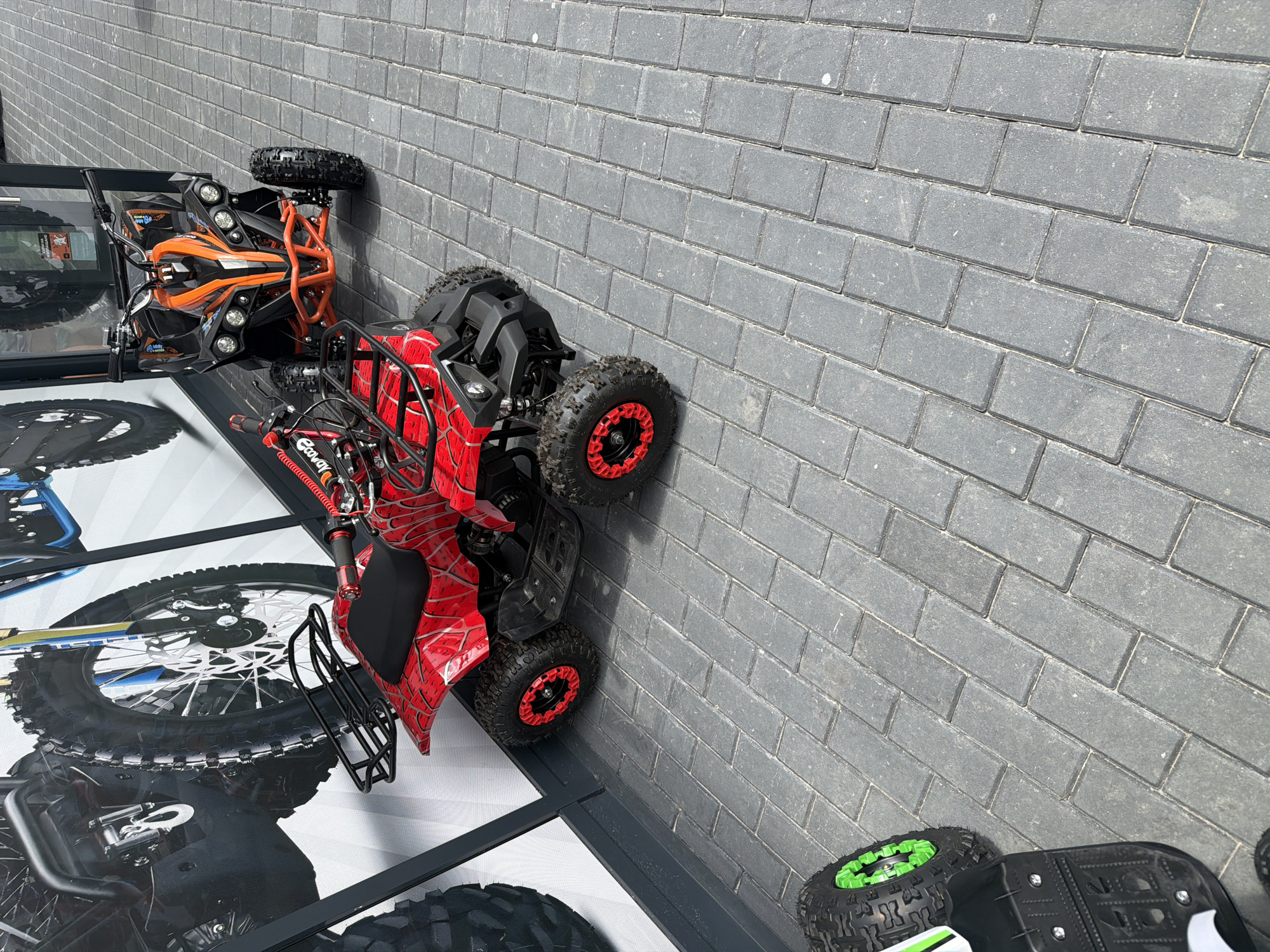 A red all-terrain vehicle (ATV) with a Spider-Man themed design is parked on a paved walkway in front of a store. Another ATV, orange and black, is parked to its right. The ATVs appear to be child-sized. In the background, a large graphic of a motorcycle is displayed on the wall of the building. The walkway is made of gray rectangular pavers. The scene is well-lit, suggesting daytime. This image was taken in Rakovski, Bulgaria.