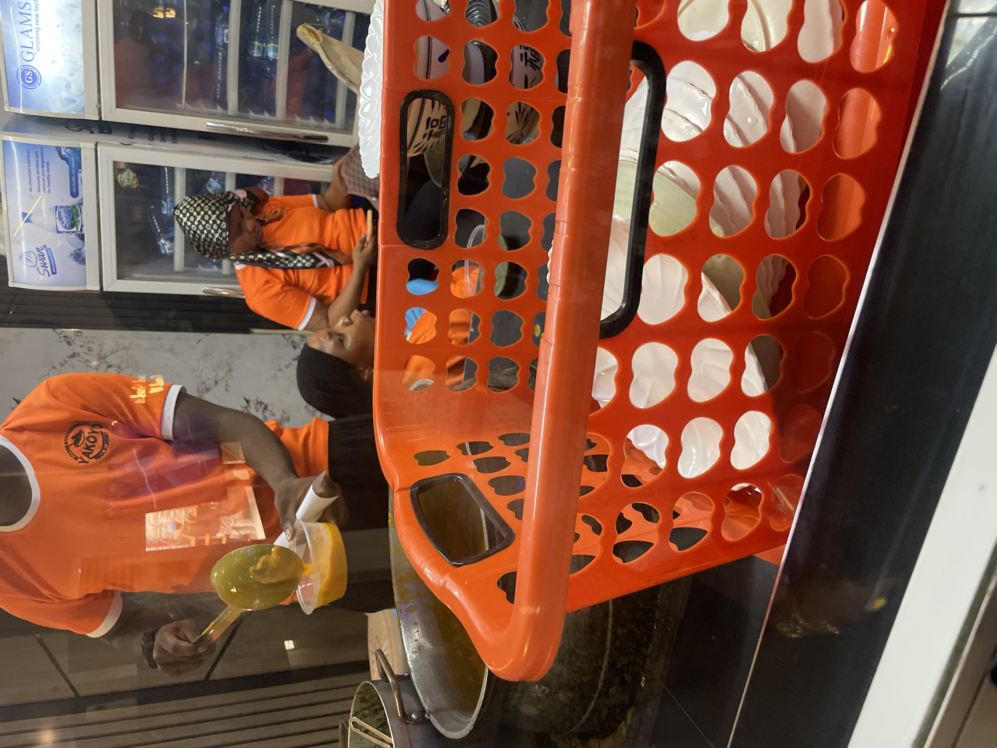 In Abuja, Nigeria, a woman wearing a black and white head covering and an orange shirt stands in front of a cooler filled with water bottles. She is interacting with another person wearing an orange "YAKOYO" t-shirt, who appears to be pouring a yellow-green substance from a clear container with a yellow lid using a spoon. An orange shopping basket is in the foreground.