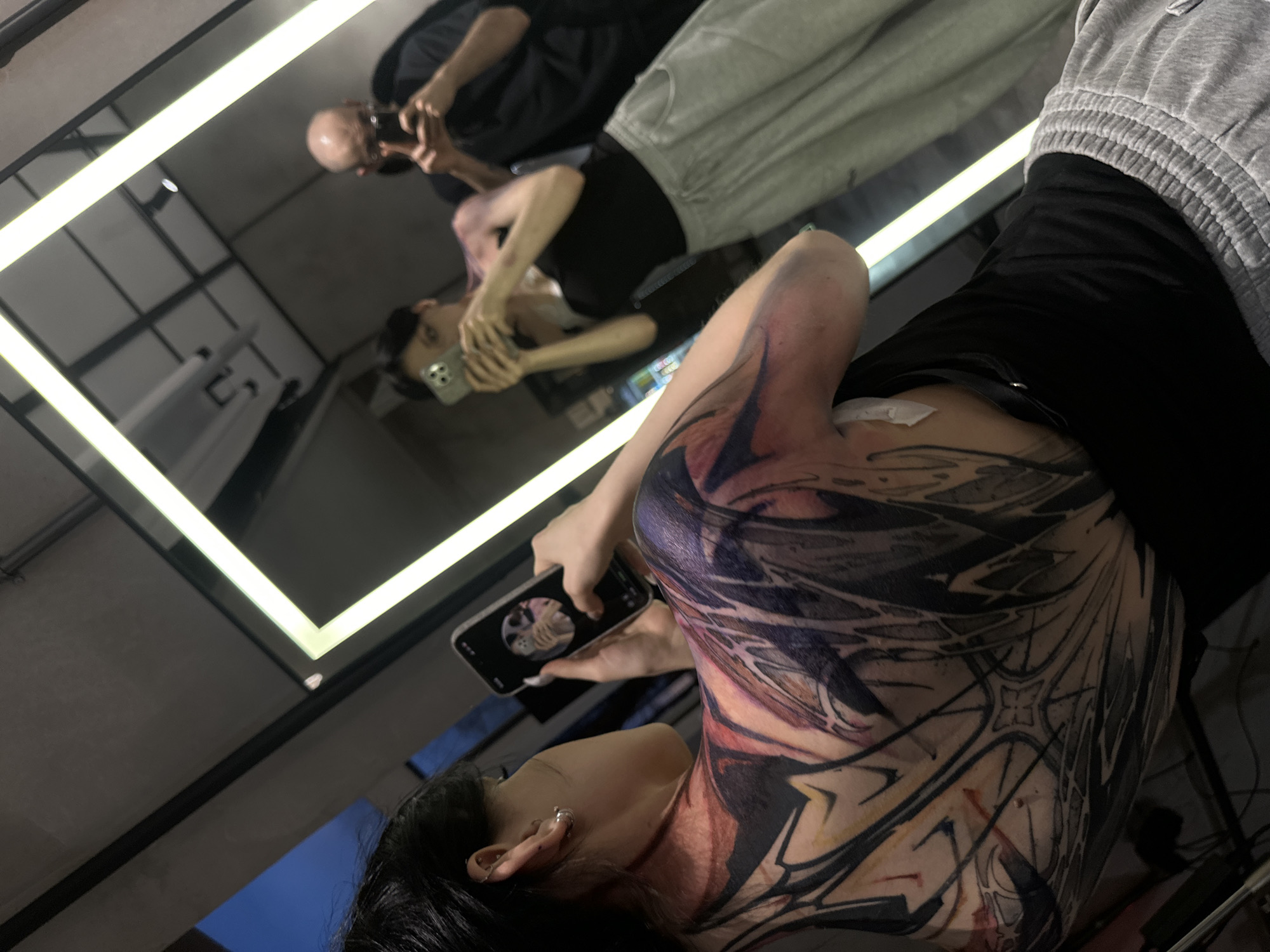 The image shows a person with a large tattoo on their back and upper arm, taking a photo of themselves in a mirror. The tattoo is primarily black with some red and purple shading, featuring complex geometric and abstract designs. The person is holding a black smartphone in their right hand. Reflected in the mirror are two people: one taking the photograph and one older person holding a phone. The environment appears to be a studio or apartment, possibly in Moscow, Russia.