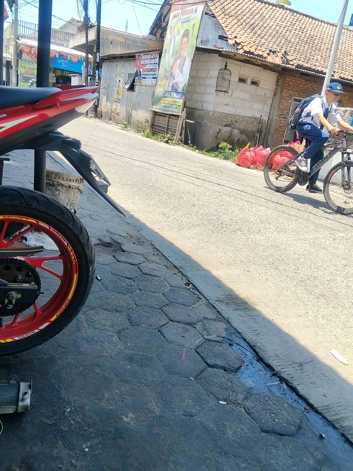 This is a medium-angle, eye-level shot taken outdoors on a street in Mekarmukti, Indonesia. A person wearing a blue cap and school uniform is riding a bicycle on the asphalt road. The cyclist is looking to their right and has a backpack on. In the foreground, the rear wheel of a red motorcycle is visible. The road surface is a mix of asphalt and paving stones. To the left of the road, there is a paved area with debris, and a small radiator is visible. To the right of the road, there are several red plastic bags. In the background, there are buildings with tiled roofs, power lines, and posters on the walls. The sky is clear and blue. The lighting suggests it is daytime.