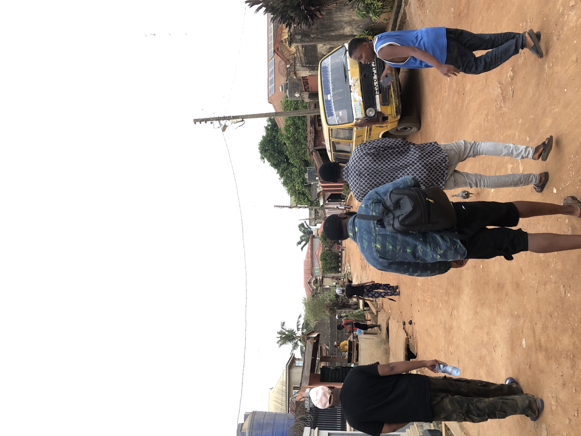 The image captures an outdoor scene featuring several men on a reddish-brown dirt road in what appears to be a semi-urban or rural setting. The perspective is from behind a group of people, looking forward down the street.
In the immediate foreground, a man wearing a black t-shirt and camouflage-patterned trousers holds a light blue water bottle. A large blue water tank is partially visible to his left. Further ahead, another man in a blue and green patterned shirt, black shorts, and a black backpack walks. Adjacent to him is a man dressed in a white patterned long-sleeved shirt and light grey trousers, with a small, dark bird visible on the ground near his feet.
Towards the mid-ground, a bright yellow minivan or bus, possibly a public transport vehicle, is positioned on the road. Its front grill and windshield are visible, displaying text like "BOBO" and what appears to be "TOYOTA" on the grille. Standing near the front of this vehicle is a man in a blue sleeveless top and dark trousers, holding a mobile phone. Behind the bus, a building features several solar panels on its roof.
The background includes various buildings with corrugated iron roofs, lush green trees, and utility poles with overhead wires lining the street. The sky is bright white, suggesting either an overcast day or overexposure. The scene portrays everyday activity on an unpaved street, with people and a vehicle.