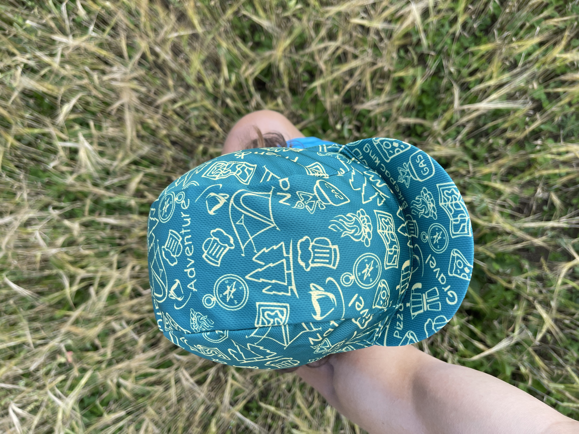 A close-up, overhead shot shows a teal cycling cap with a repeating pattern of illustrations and words, held in a person's hand. The cap features various icons related to outdoor activities and food, such as tents, campfires, coffee cups, beer mugs, compasses, pizza slices, a moka pot, and the words "Adventure," "Gravel," "Bike packing," and "GAS." The background is out of focus, showing dry grass and some green foliage, suggesting an outdoor setting. The image does not contain any information about Maksimovo, Russia.