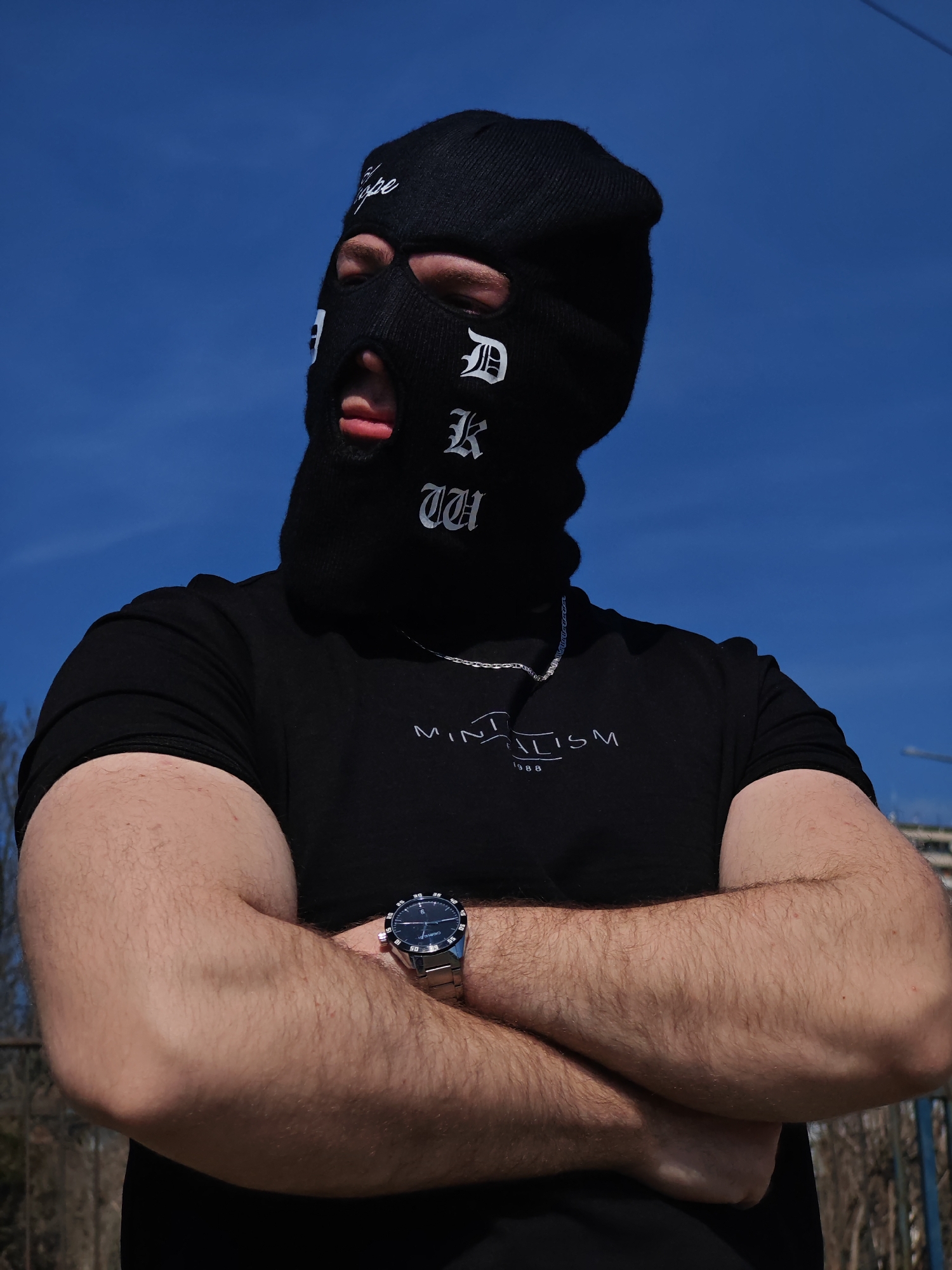 An individual is depicted outdoors against a clear, bright blue sky, likely in Shumen, Bulgaria, specifically within a general urban or residential area, evidenced by distant buildings and sparse vegetation in the background. The subject, a person with muscular, hairy forearms and biceps, is wearing a black short-sleeved t-shirt with "MINIMALISM 1988" printed in white, a silver-colored chain necklace, and a silver-colored wristwatch with a dark face on the left wrist. The head is covered by a black balaclava featuring white text: "Dope" on the forehead and "DK UH" vertically aligned on the right side of the face in a Gothic font. The individual's light-colored eyes are visible through the balaclava's eye openings, and the tongue is slightly protruding through the mouth opening. The person stands with arms crossed over the chest. Faint outlines of bare trees or bushes and a dark fence-like structure are discernible in the lower background.