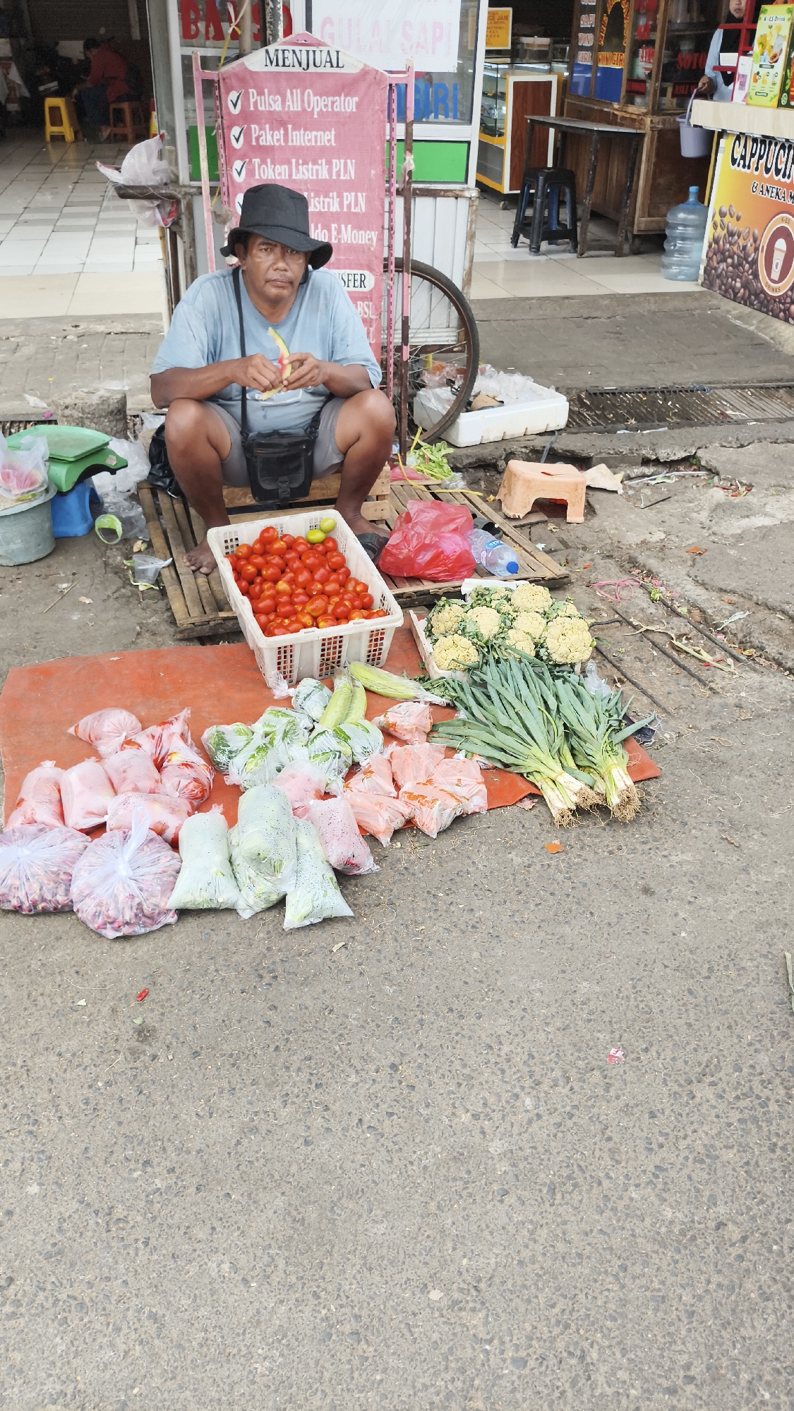 A man wearing a light blue t-shirt and a black hat is crouching down in front of a stall. He is holding a piece of fruit, possibly a banana, and appears to be peeling it. In front of him, on a red tarp, are several bags of produce, including tomatoes, cauliflower, leeks, and other vegetables. Behind him, a sign lists various services, including phone credit, internet packages, and electricity tokens. The scene appears to be an outdoor market, possibly in South Jakarta, Indonesia, with buildings and other stalls visible in the background. The man is the central subject of the image, and his activity suggests he is taking a break or preparing food. The surrounding produce indicates a marketplace setting.