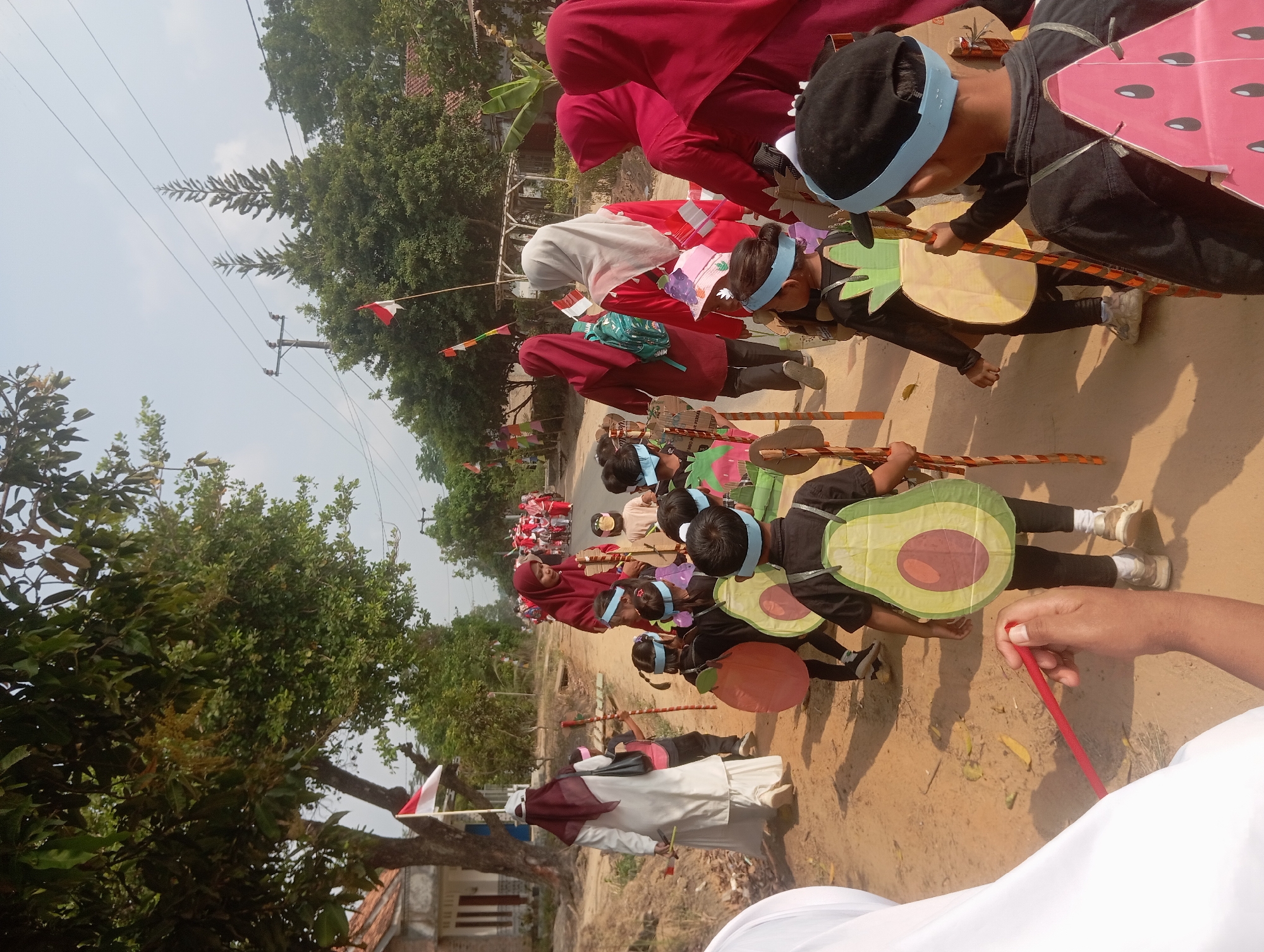 This image captures a festive parade in Dente Teladas, Indonesia, likely celebrating a national holiday. A group of children, dressed in colorful fruit-themed costumes, walk down a dirt road. Their outfits include large, cut-out representations of avocados, pineapples, and watermelons worn as backpacks. They are accompanied by adults, some wearing hijabs and traditional clothing, who appear to be chaperoning or participating in the event.
The environment is a rural setting with a dirt road lined by trees and vegetation. In the background, there are houses and power lines, typical of Indonesian villages. Indonesian flags are prominently displayed on poles and attached to some of the participants, indicating a patriotic celebration. The weather appears sunny and clear. The children are holding wooden poles, some decorated with colorful ribbons, which they likely use to support their costumes or as part of the parade's activities. The overall scene depicts a lively community event, possibly a school parade or a local festival.