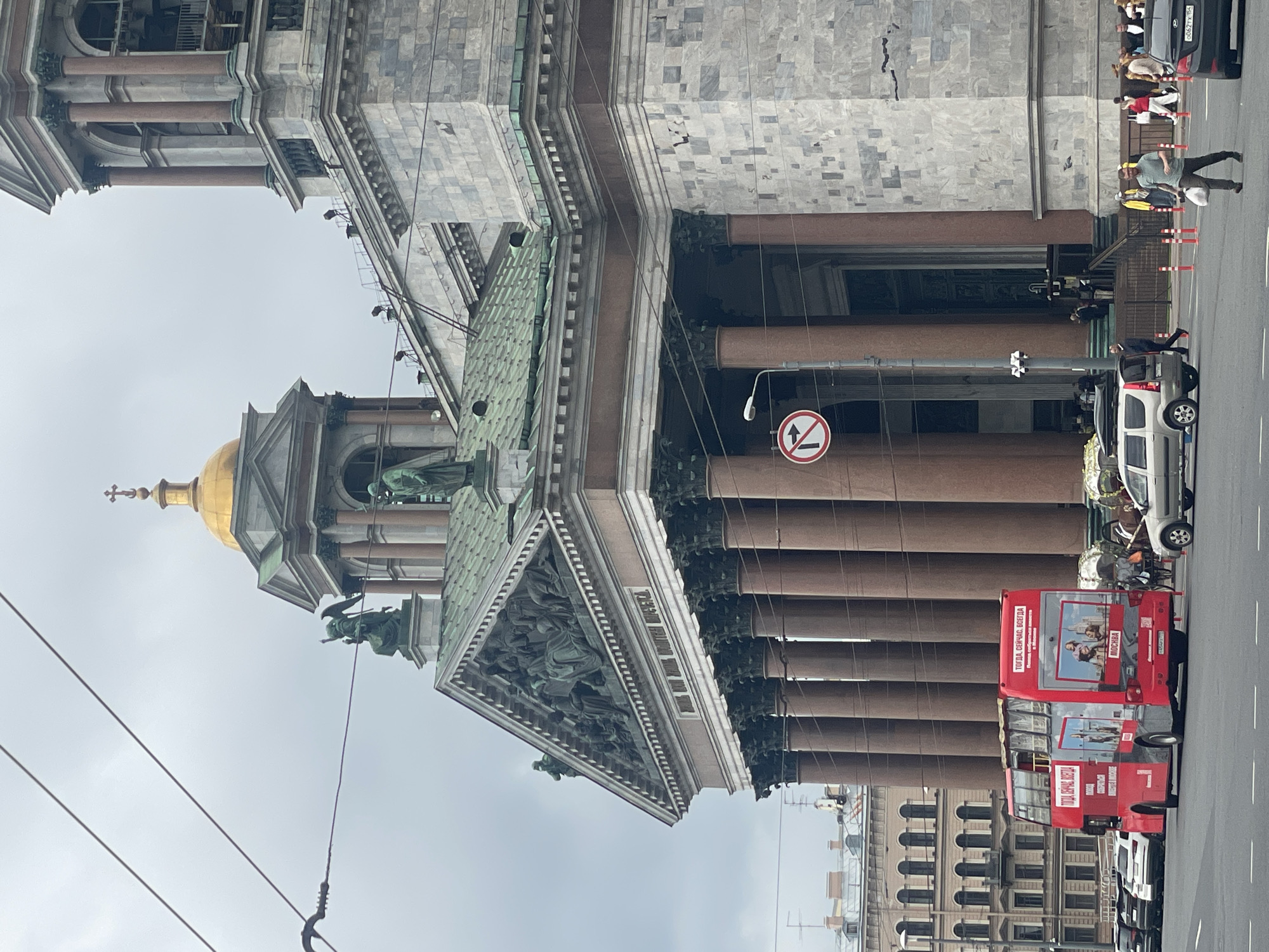 The image captures a view of St. Isaac's Cathedral in Saint Petersburg, Russia, on an overcast day. In the foreground, a red double-decker tour bus is parked on the left side of the street. Several cars are also visible on the street, including a silver SUV to the right of the bus. Pedestrians are walking on the sidewalk in front of the cathedral.
St. Isaac's Cathedral dominates the background, with its massive granite columns, gilded dome, and intricate architectural details. The façade features sculptures and relief carvings. A traffic sign indicating no right turn is posted on a lamppost. The sky is cloudy, suggesting it is either early morning, late afternoon, or a generally overcast day. The overall scene depicts a bustling urban environment with iconic architecture.