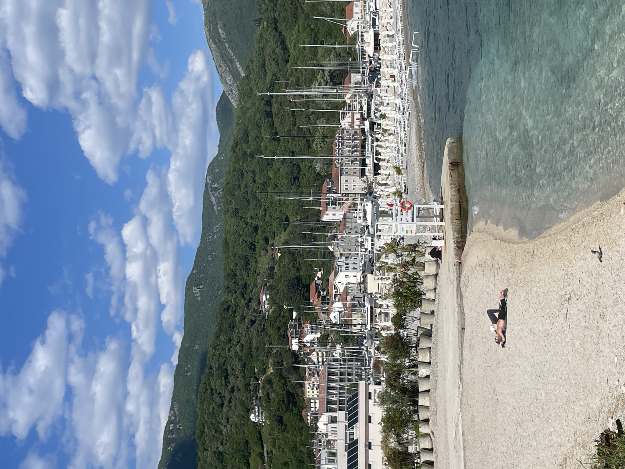 This media file captures a picturesque coastal scene in Herceg Novi, Montenegro, from a slightly elevated vantage point overlooking a bay. The image is bathed in bright daylight under a clear blue sky dotted with scattered white clouds.
In the foreground, a light-colored sandy or concrete promenade runs along the water's edge. A person is seen lying on their back on this surface, relaxing in the sun, dressed in dark shorts and a light top. Another person is partially visible sitting further down the promenade. A small bird is also present on the ground. To the left, a series of light-colored, low concrete block-like structures line the edge of the promenade, behind which are some green bushes. A red and white lifebuoy is attached to a white structure near the water.
The clear, turquoise-green water of the bay is visible to the right, appearing calm and inviting. Along the shoreline, particularly to the left of the water, numerous white sun loungers are neatly arranged, indicating a beach or resort area.
Beyond the promenade and beachfront lies a bustling marina filled with numerous sailboats and yachts, their tall masts creating a dense, vertical pattern against the backdrop. Residential and commercial buildings with varied architectural styles, including multi-story structures with tiled roofs, are nestled amongst the boats and climb the lower slopes of the mountains.
The background is dominated by majestic, steep mountains covered in dense green foliage, stretching across the horizon and providing a dramatic natural setting for the town. The overall impression is one of a vibrant and tranquil Mediterranean coastal town, ideal for leisure and boating activities. There is no visible text.