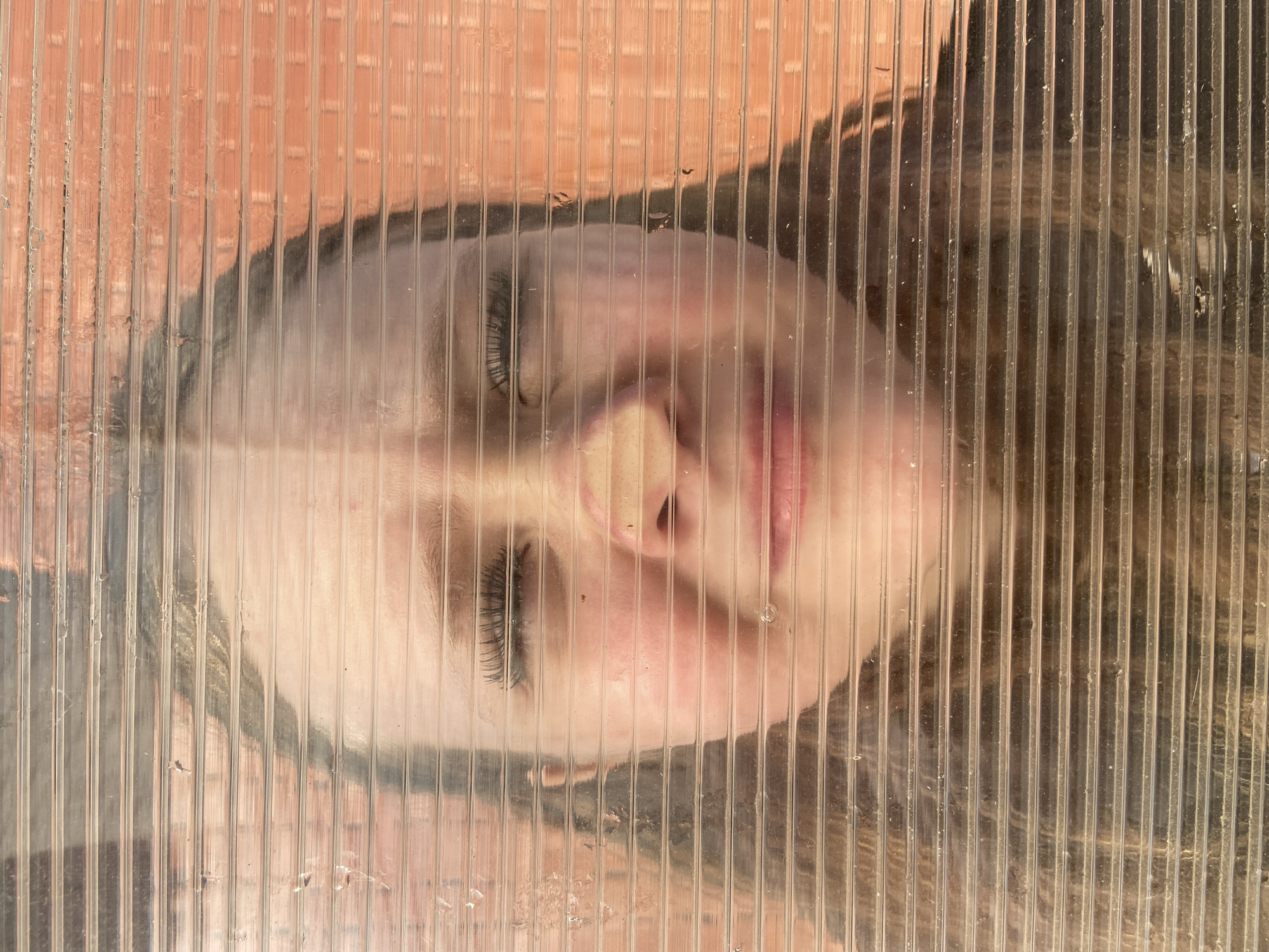 The image shows a woman's face pressed against a translucent, ridged plastic or glass surface, possibly a window. Her eyes are closed, and her expression is serene. The light is soft and even, casting a warm glow. The background is obscured by the translucent material, but a brick structure is visible. The specific location in Chaikovsky, Russia, is not evident from the image.