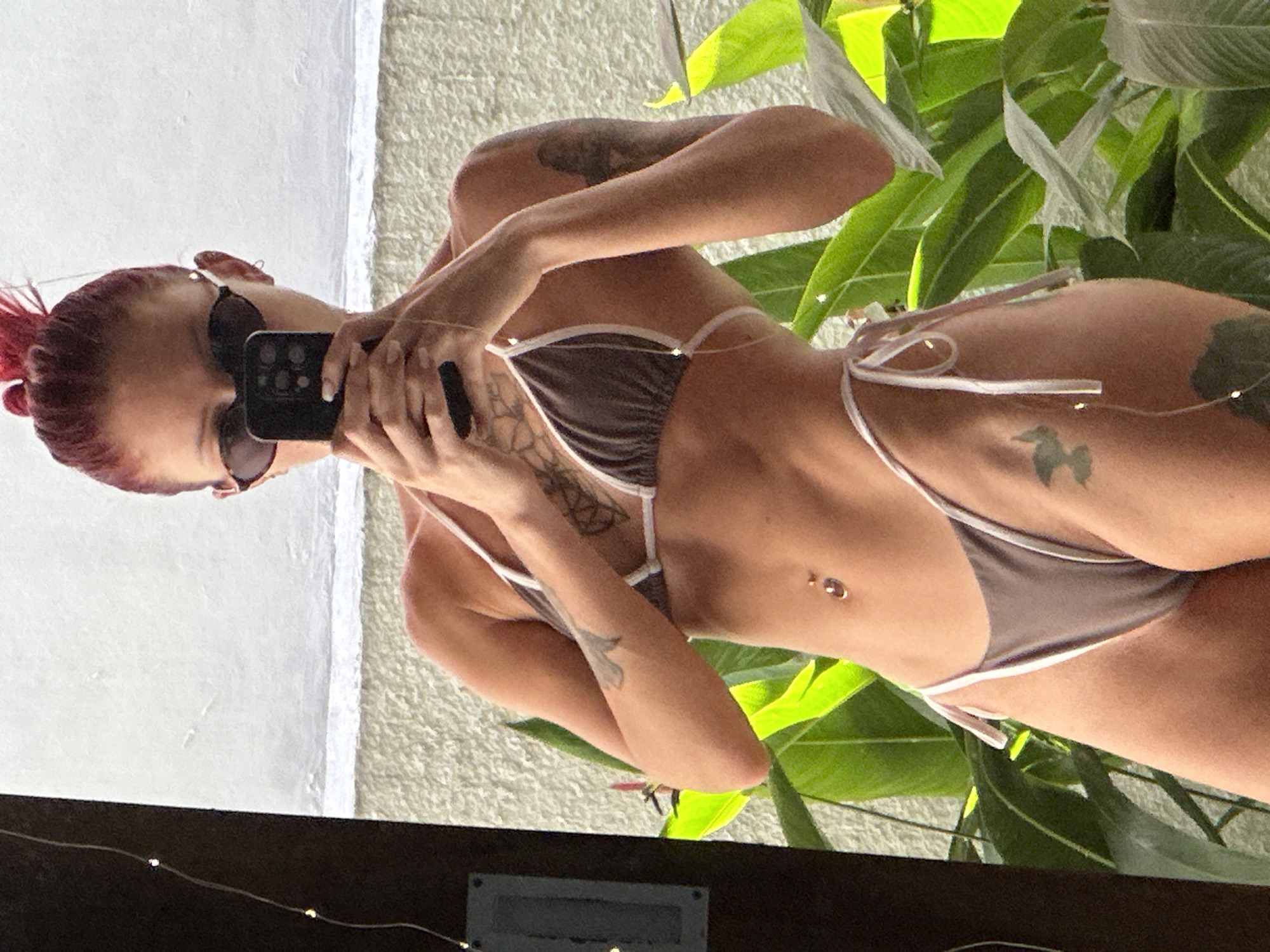 A woman is taking a selfie in a mirror. She is wearing a brown bikini with white trim. She has red hair pulled back in a bun and is wearing sunglasses. She has several tattoos on her arms and torso. The background includes a white wall, a textured rug, and green plants. A dark brown surface with small lights is visible at the bottom of the image, reflecting the scene. There is no information present to verify the location as the city of Dalung, Indonesia.