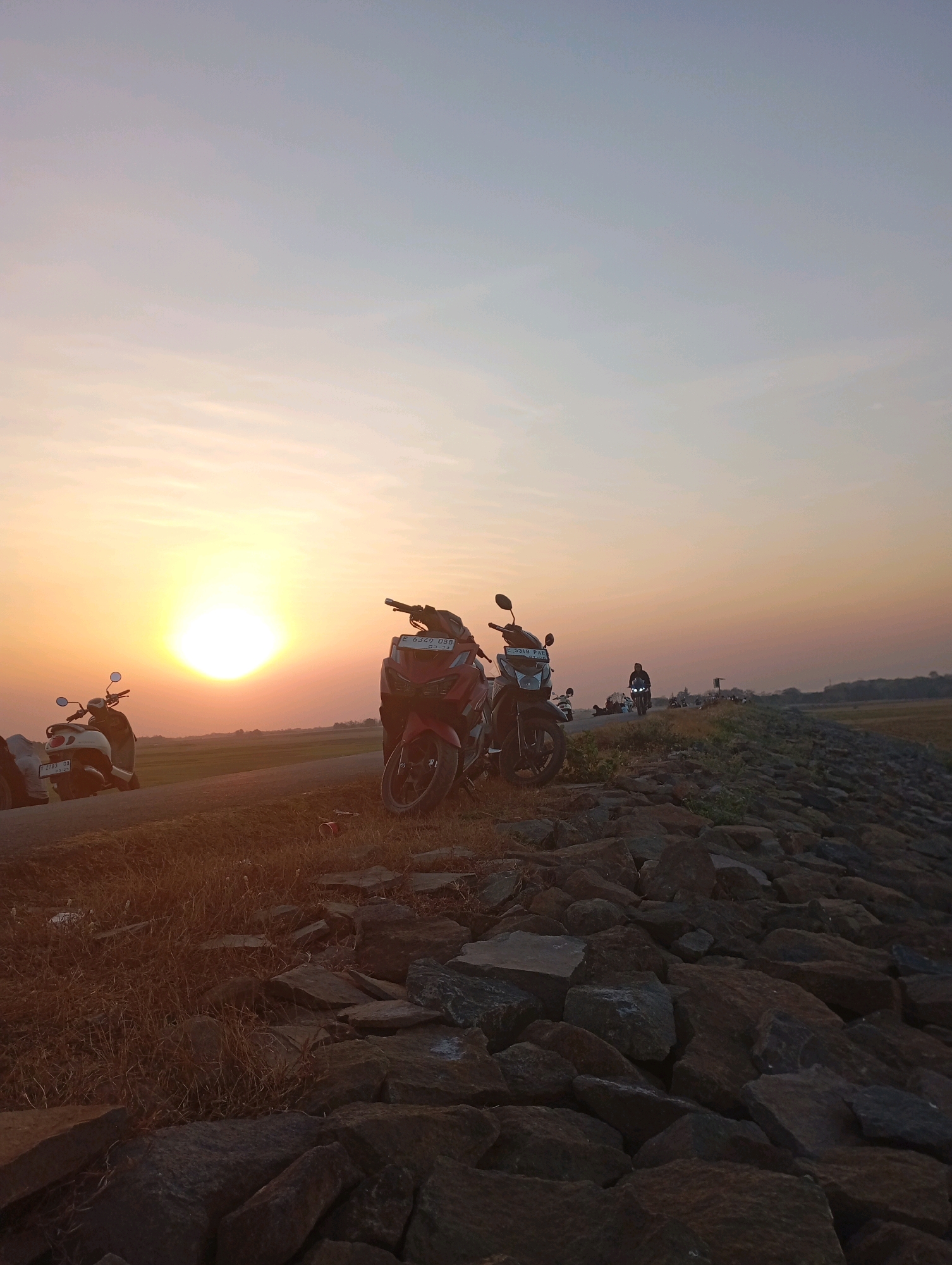 This image captures a scenic sunset at the Haurkolot dam in Indonesia. The sky transitions from a soft orange near the horizon to a pale blue overhead, with the sun prominently positioned on the left, casting a warm glow. The foreground is dominated by a rocky embankment, part of the dam's structure, covered in scattered stones and dry grass. To the left of the embankment, a paved surface is visible, where several motorcycles are parked. One red scooter and one white scooter are clearly visible. Further down the paved area, a group of people and more motorcycles are present, with one individual riding a motorcycle towards the viewer. In the background, a treeline is visible against the horizon, indicating the surrounding landscape. The overall scene suggests a gathering of people enjoying the evening by the dam.