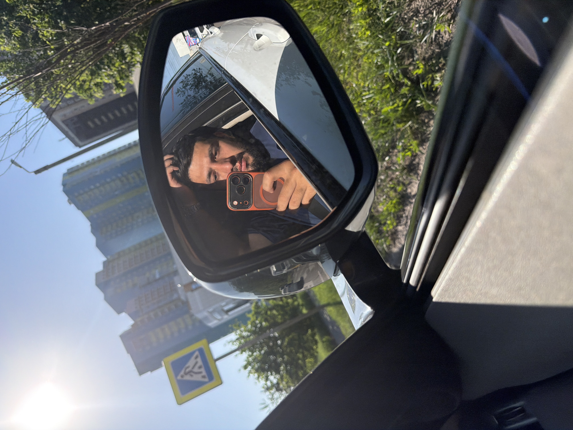 The media file depicts a mirror selfie taken by a man from inside a car. The man, featuring dark hair and a beard, is prominently reflected in the car's side-view mirror. He is holding an orange-cased smartphone, which displays multiple camera lenses, to capture his own image in the mirror. His tongue is playfully extended, suggesting a casual or humorous expression.
The bright daylight causes a strong sun flare in the lower left of the frame. Beyond the car and its mirror, a clear blue sky dominates, interspersed with green foliage from trees and bushes. In the background, modern, multi-story buildings with numerous windows are visible against the sky, one appearing yellow/gold and another grey/silver. A blue square pedestrian crossing sign with a yellow border and a white symbolic figure is also visible, appearing inverted due to the image's overall rotation.
Part of the car's white door is reflected in the mirror and visible outside its frame, alongside the dark interior trim of the car's window. The scene captures a sunny day in an urban environment, consistent with the provided location context of Krasnoyarsk, Russia.