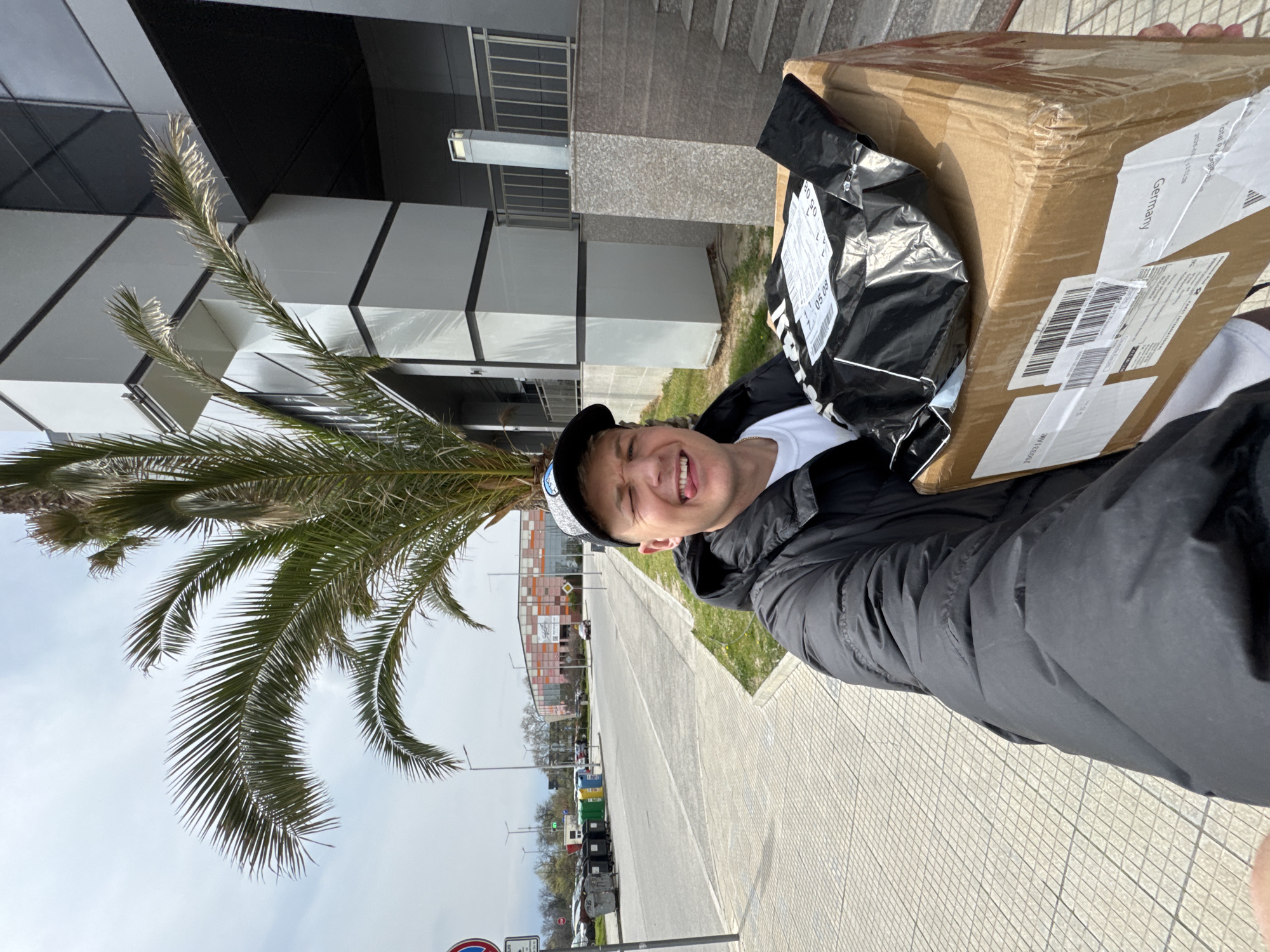 A man with blond hair and a black cap stands on a sidewalk, holding a large cardboard box with shipping labels, one of which reads "Germany." He is wearing a white t-shirt and a black jacket. The man is smiling and sticking out his tongue. Behind him, there is a modern building with black and white geometric patterns, a palm tree, and a street with recycling bins and parked cars. In the background, there is a building with a colorful exterior. The location is Plovdiv, Bulgaria.
