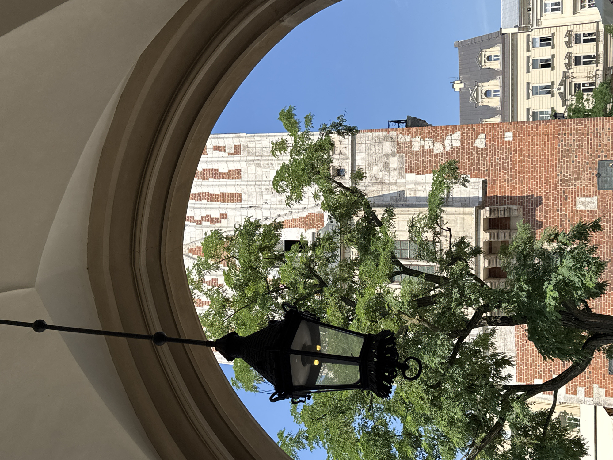 This is a ground-level, eye-level, outdoor, medium shot photograph taken on a clear day in Krakow, Poland, likely in the Main Market Square. The image is framed by a large, beige, arched stone structure, possibly part of a building or portico. Hanging from the archway is a black, ornate, wrought-iron lantern with glass panes, illuminated by two visible yellow bulbs.
In the background, through the arch, a tall, multi-story tower with red brick and white stone detailing is visible. The tower has a distinct architectural style with decorative stone elements and arched windows. To the right of the tower, a lighter-colored building with a grey mansard roof and dormer windows is seen. The lower part of this building features multiple windows and decorative architectural elements.
A lush green tree with abundant foliage partially obscures the tower and the buildings. The leaves are vibrant and indicate a season of growth. The sky is a clear, bright blue. The overall impression is of a historical urban setting, possibly a public square or courtyard, bathed in sunlight. There are no people or animals visible in the frame.