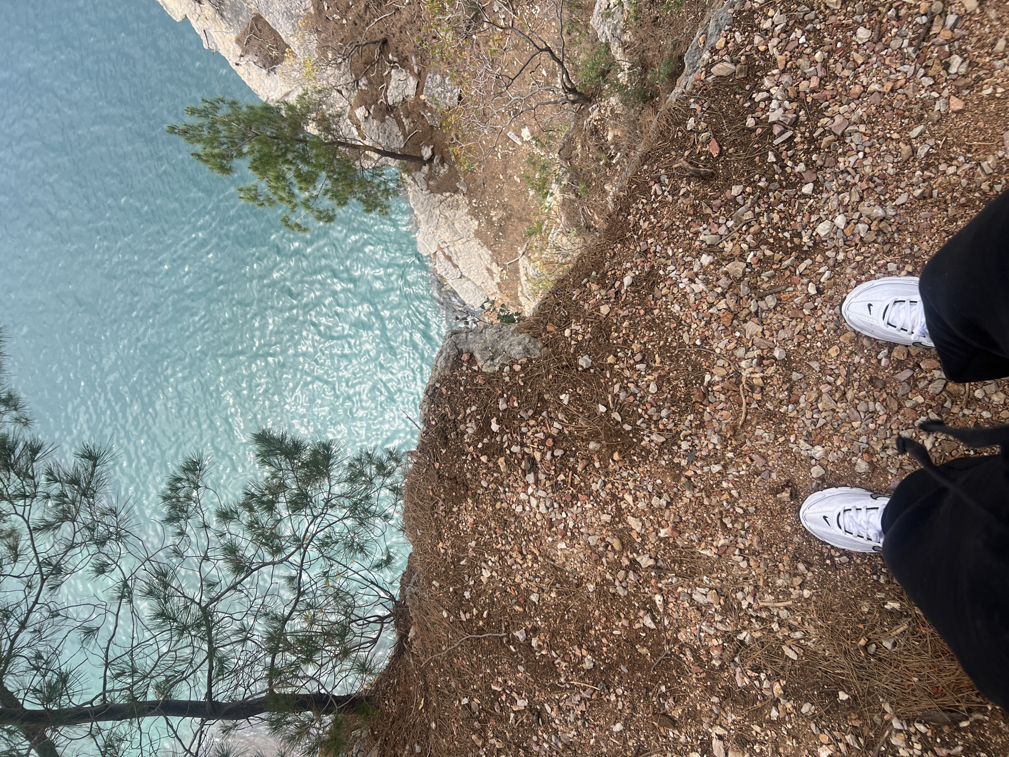 The image shows a view from the edge of a cliff in Petrovac centar, Montenegro, looking down at turquoise water. The ground at the edge of the cliff consists of rocks, gravel, and dead leaves. A person wearing white sneakers and black pants stands at the edge. Several pine trees grow on the side of the cliff.