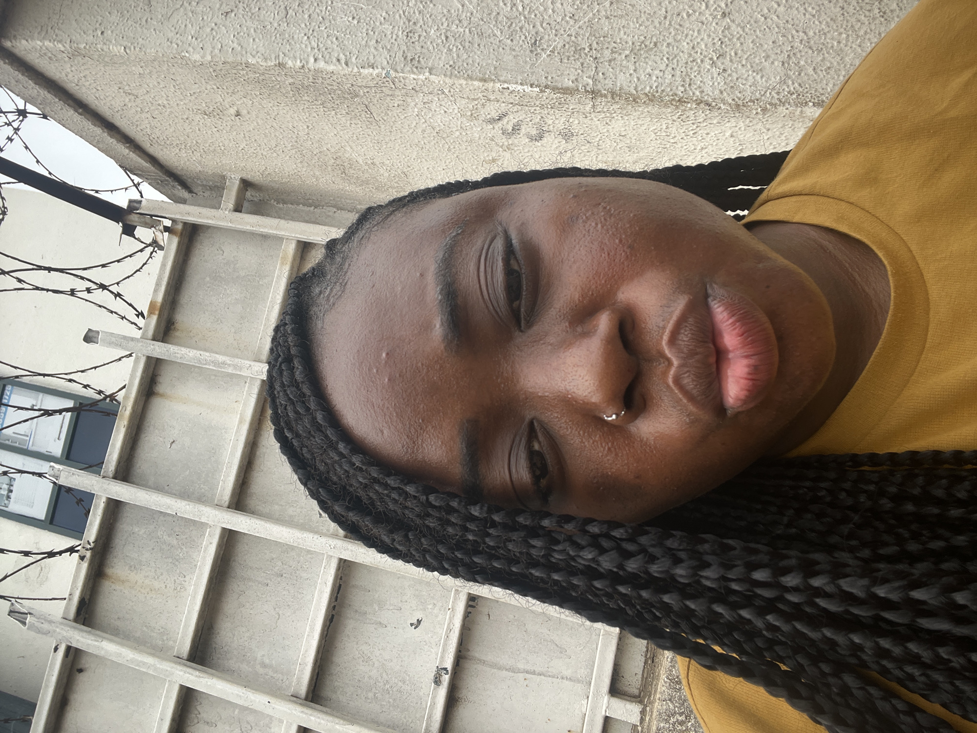 The image is a close-up portrait of a dark-skinned individual, likely female, captured outdoors. The person's face and upper body are prominent, with their head significantly tilted towards the bottom right of the frame. They are looking directly at the camera with a neutral to slightly relaxed expression, their lips slightly parted. A small silver stud nose piercing is visible on their right nostril. Their hair is styled in long, dark box braids, some of which fall over their shoulder. The person is wearing a mustard yellow top or t-shirt.
The background features a light-colored, textured concrete or stucco wall, which due to the image's rotation, appears above and to the left of the person's head. A metallic structure, resembling a fence or gate with horizontal and vertical bars, is visible behind the person's head and hair. Strands of barbed wire are coiled along the top section of this metallic structure, particularly in the upper left portion of the frame. Through the gaps in the fence and barbed wire, a window with dark frames is partially discernible. Faint, dark numbers or letters, possibly "238 1/25", are visible on a light-colored surface inside the window. The sky, visible in the very top-left corner, appears overcast, indicating daytime. The setting suggests an urban outdoor environment, potentially in Abuja, Nigeria, featuring a building and security fencing.