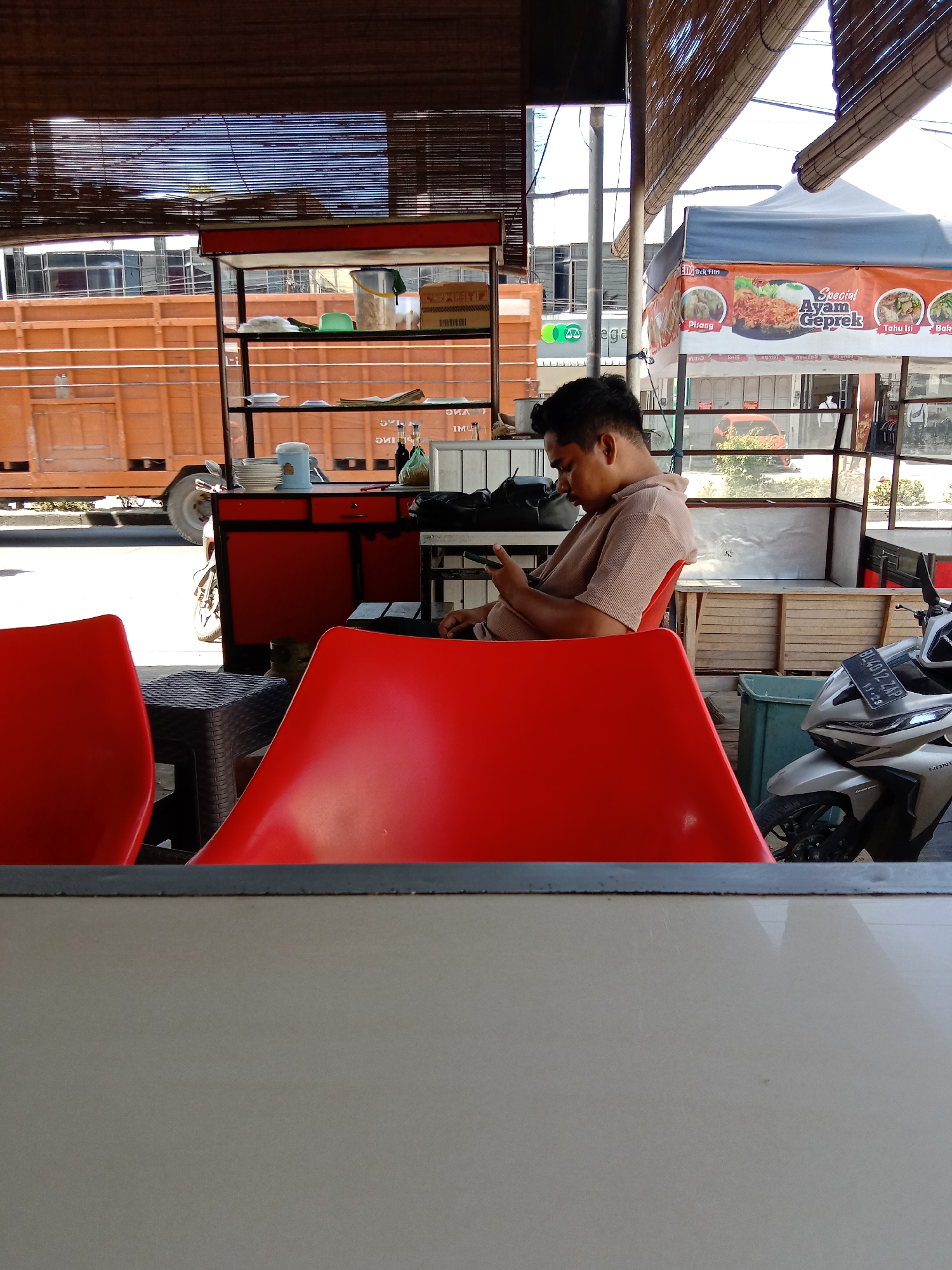 In Lhokseumawe, Indonesia, a man sits at a red plastic chair with his phone, under a bamboo awning. He is wearing a tan, striped, long-sleeved shirt. In front of him, another red chair and a dark plastic stool are visible. Behind the man, a food stall with a tent displaying "Special Ayam Geprek" and other food items is set up. A silver motorcycle with a license plate is parked to the right of the stall. In the background, a large orange truck is parked on the side of a street with buildings in the distance.