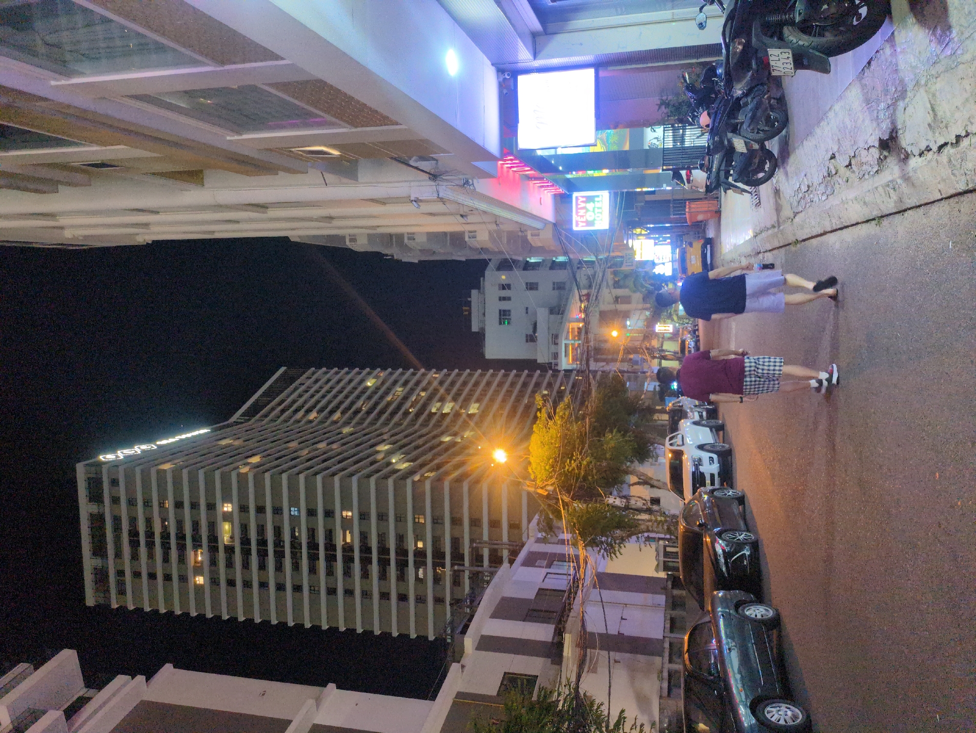Captured at night in South Quy Nhon Ward, Vietnam, the image reveals a street scene with pedestrians walking along the road, and parked vehicles. Towering buildings illuminate the urban landscape. Street-level commerce and signage are visible among the motorbikes parked along the curb.