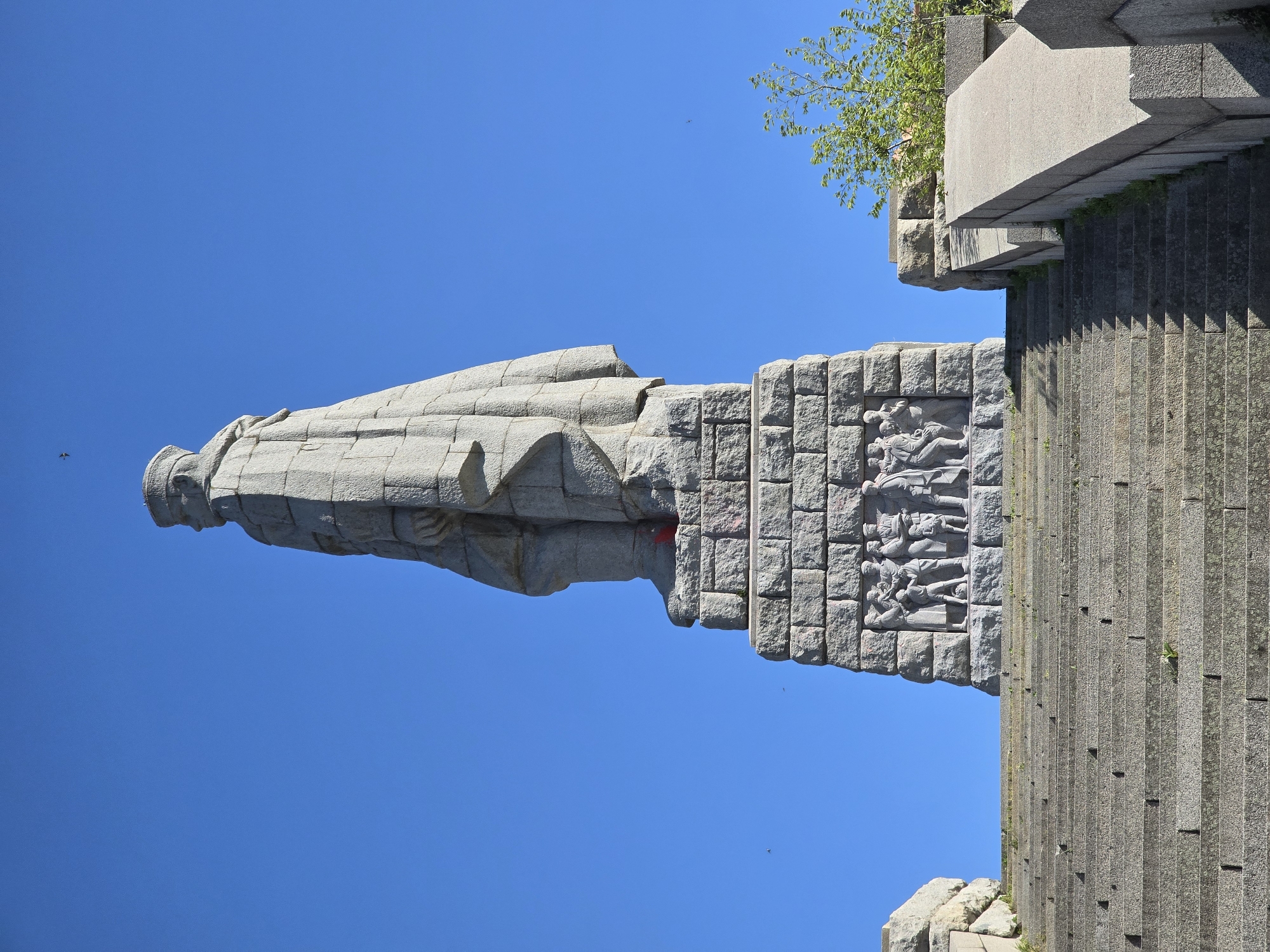 The image shows the Monument of the Soviet Army in Plovdiv, Bulgaria. The monument consists of a large, stone figure of a Soviet soldier, standing atop a tall pedestal. The pedestal is adorned with sculpted reliefs depicting scenes of Soviet soldiers interacting with civilians. The monument is located on a hillside, with a long set of stone steps leading up to it. The sky is clear and blue.