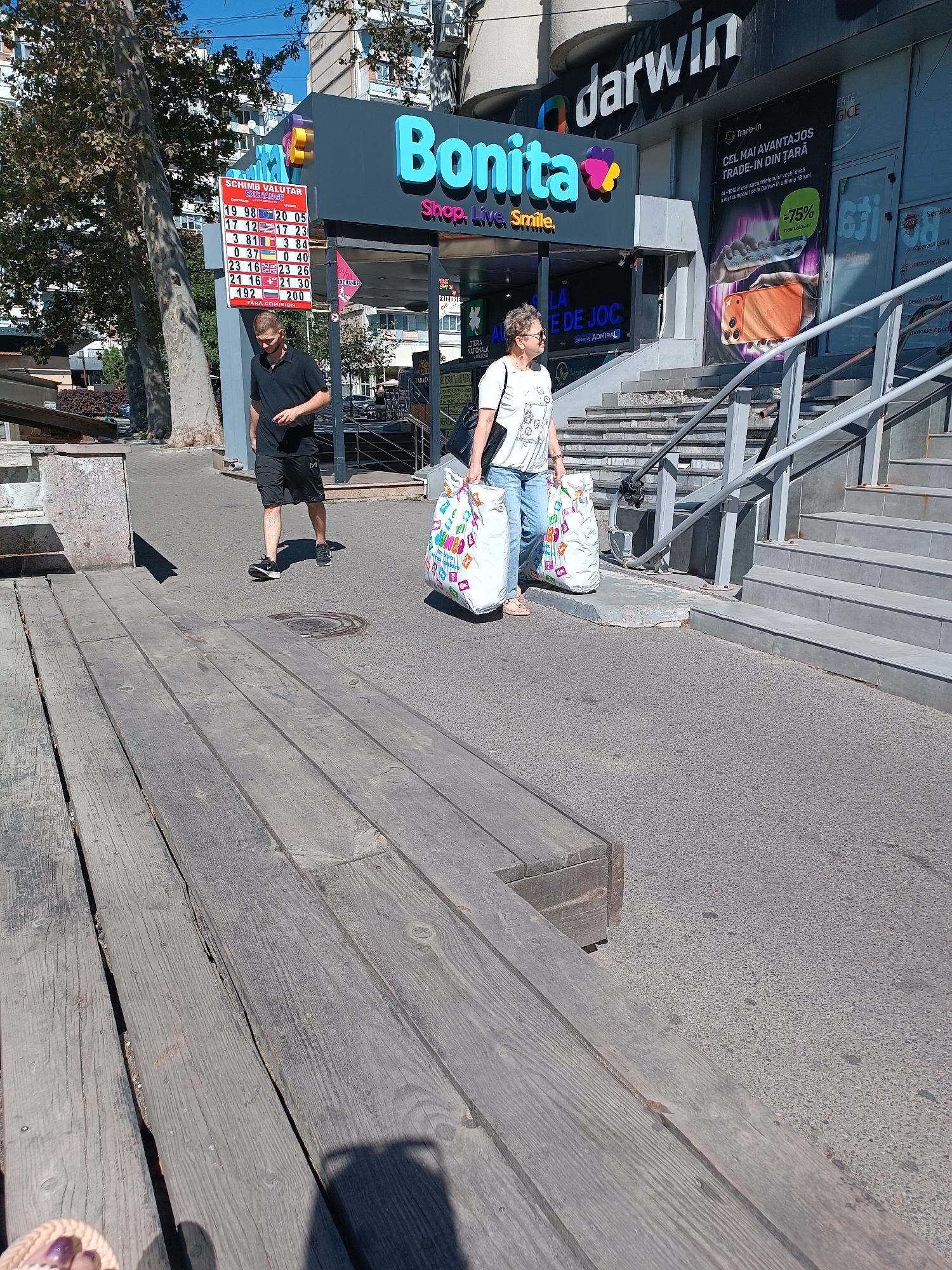 The image depicts a street scene in Chișinău, Moldova, on a sunny day. In the foreground, a wooden walkway runs parallel to the street. On the street, a man wearing a black polo shirt and shorts walks away from the camera, while a woman wearing a white t-shirt and jeans walks towards the camera, carrying two large shopping bags.
Behind the pedestrians, a "Bonita" shop is visible, featuring a bright, colorful sign. Adjacent to it, a "Darwin" store, specializing in electronics, is advertised with a poster promoting a trade-in service for phones, highlighting a "75% value" offer. Further down the street, a currency exchange board displays exchange rates for various currencies, including USD, EUR, and RUB. The environment appears to be an urban commercial area with a clear blue sky overhead.