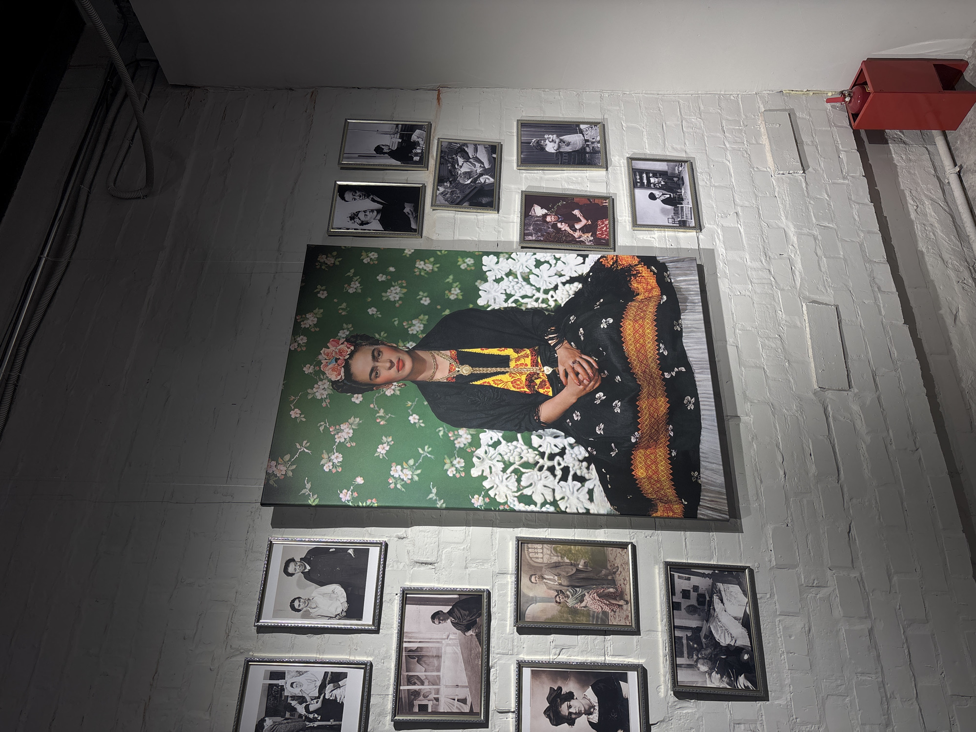 The image shows a white brick wall adorned with a large portrait of Frida Kahlo surrounded by smaller framed black and white photographs. The portrait features Kahlo wearing a black top with an ornate gold and yellow necklace, lying against a green background with white floral patterns. Her head is adorned with flowers. Above the portrait are several framed photographs, seemingly arranged in a cluster. Below the portrait are more framed photographs. A red fire extinguisher box is mounted on the wall in the upper right corner of the frame. The light source appears to be from the top, casting shadows on the wall. There is a dark area in the top left corner, possibly indicating a ceiling or corner of the room. The location is unknown and there is no clear evidence it is Krasnoyarsk, Russia.