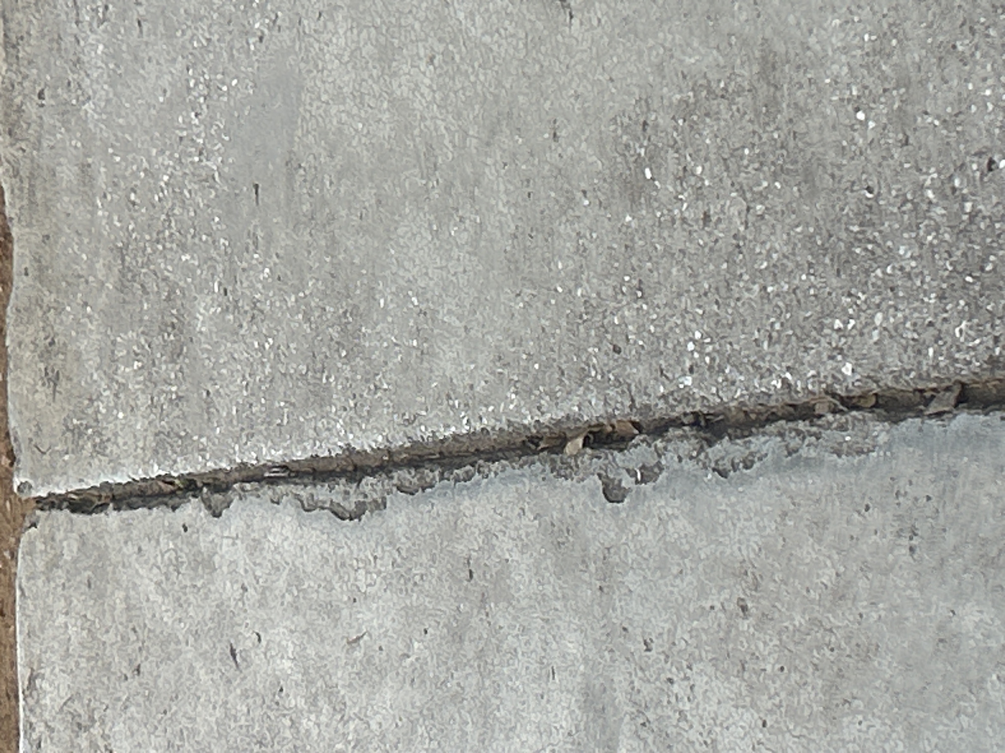 The image shows a close-up view of a crack in a concrete surface. The crack runs vertically through the center of the image, dividing the concrete into two sections. The concrete appears aged and weathered, with small pebbles and debris embedded in its surface. The crack itself is irregular in shape and appears to be deep, with some organic material, possibly leaves or dirt, visible within it. The lighting suggests an outdoor environment, likely during the daytime. There are no people or animals visible in the image. The scene is devoid of any specific identifiable landmarks that would indicate its location within Santa Ana, Venezuela.