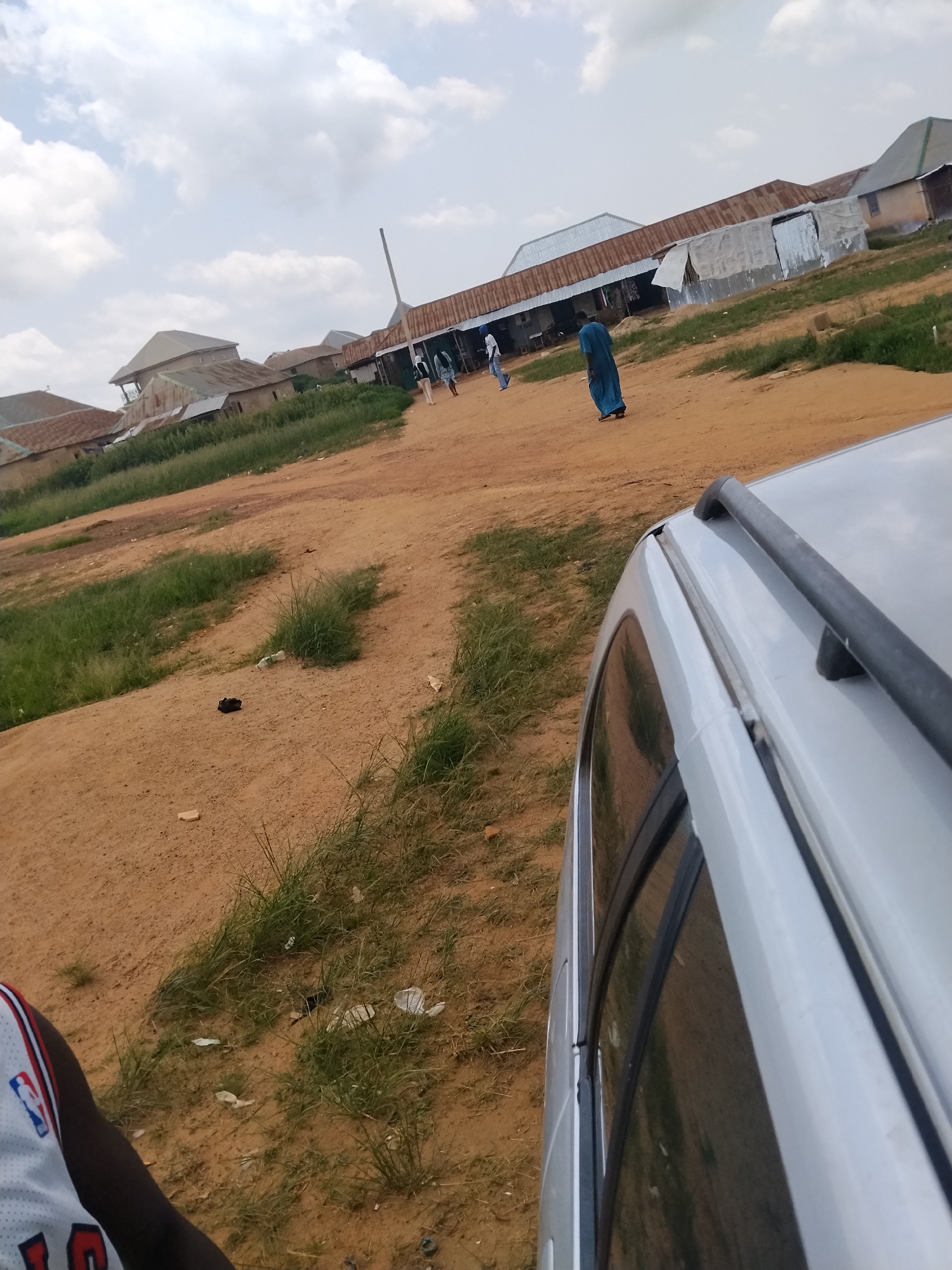 The image shows an outdoor scene in Durumi, Nigeria, under a cloudy sky, suggesting it might be mid-day or late afternoon. In the foreground, the edge of a silver car is visible, with its roof rack and part of the window. To the left of the car, a person wearing a white jersey with red and blue trim is partially visible, facing away from the camera.
The main focus of the scene is a dusty, unpaved path leading towards a row of buildings. These buildings appear to be shops or market stalls, characterized by corrugated metal roofs, some of which are rusted. In the middle ground, a few individuals are walking along the path. One person, dressed in a long blue robe, is walking away from the camera towards the buildings. Another person in a light-colored outfit is seen walking in the same direction, further down the path. There are also other indistinct figures further in the distance.
The area around the path is covered with patches of dry grass and sparse vegetation, indicating a semi-arid or dry season environment. Scattered on the ground are a few pieces of debris, including plastic bottles and paper. The overall impression is a bustling, informal marketplace or community area in Nigeria. There is no visible text in the image.