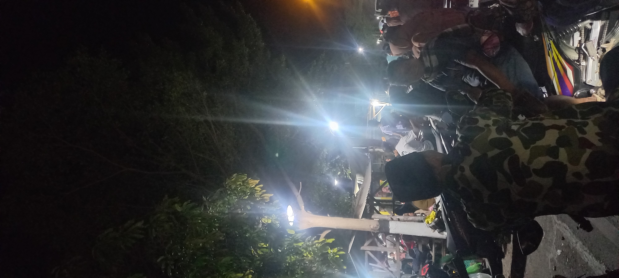 The image captures a nighttime scene in Tongaswetan, Indonesia, possibly near a roadside market or gathering. In the foreground, a person wearing a camouflage shirt and a dark cap is seated on a motorcycle. Behind them, a crowd of people is visible, some sitting and others standing. There are several motorcycles parked, and some stalls or structures suggesting a temporary setup. Bright streetlights and possibly vehicle headlights illuminate the scene, creating lens flares and casting shadows. The environment appears to be outdoors, with trees and foliage visible in the background. The overall atmosphere is one of a casual, possibly social, gathering at night.
