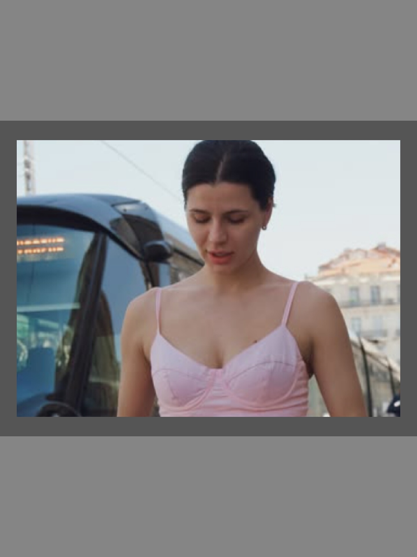 In a medium shot, a young woman with dark hair pulled back stands outside, looking down. She is wearing a pale pink, strappy top that accentuates her décolletage. To her left, the dark, curved front of a modern tram is visible, with its headlights or interior lights glowing faintly. Behind her, the background is softly blurred, showing what appears to be the facade of a building with architectural details, suggesting an urban environment. The lighting is bright and natural, indicating it is daytime.