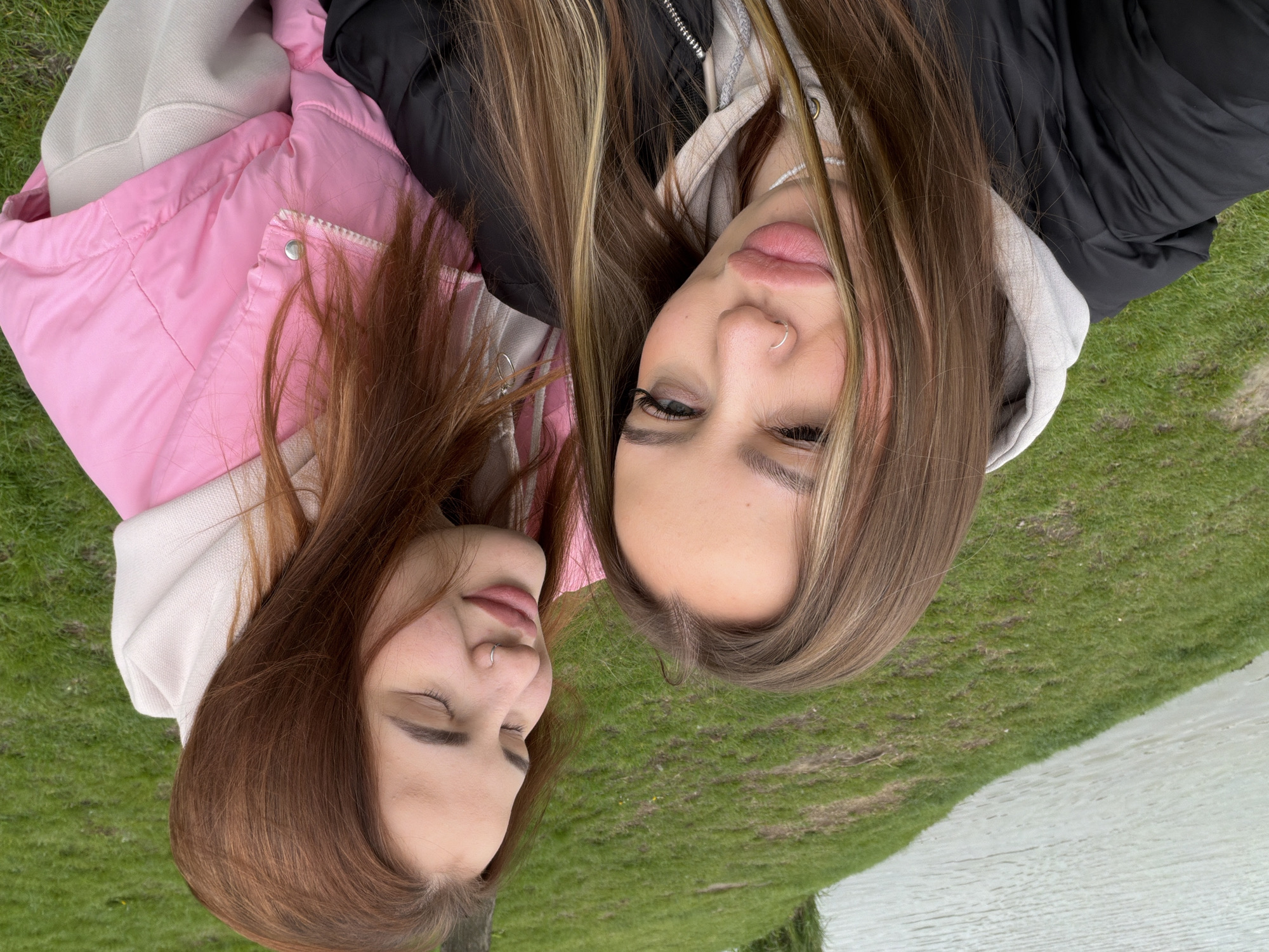 Two young women are captured in a close-up, top-down shot, appearing to lie side-by-side on green grass outdoors. The image is rotated, presenting the subjects horizontally.
The woman on the left has long, reddish-brown hair and is wearing a light pink puffer vest over a light-colored hoodie. Her eyes are closed, conveying a peaceful or serene expression, and she has a silver nose ring. Her lips are slightly parted.
The woman on the right, positioned close to her, has long, brown hair with distinct blonde highlights. She is dressed in a black puffer jacket over a light grey hoodie. Her eyes are open and directed towards the viewer, and she also wears a silver nose ring. A delicate white beaded necklace is visible around her neck, partially obscured by her hoodie. Her lips are full and slightly open.
The background consists of vivid green grass, indicating an outdoor setting, which transitions into a calm body of water, possibly a river or lake, on the right side of the frame. The lighting suggests daytime, likely overcast, with soft, diffused light. The overall mood is casual and relaxed.
The activity appears to be posing for a portrait photograph in a natural environment. The scene takes place in Krasnodar, Russia. There is no visible text in the image.