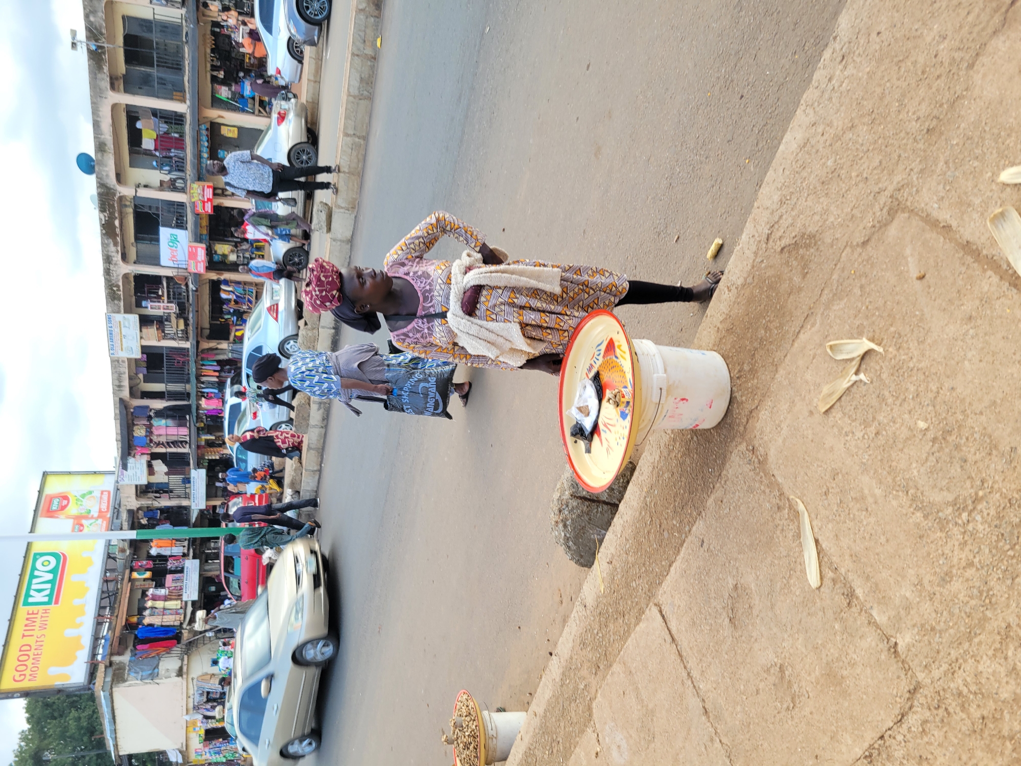 This is a medium shot, outdoor, eye-level photograph, taken in daylight. The scene depicts a bustling street market in Jos, Nigeria. In the foreground, a woman wearing a colorful patterned dress and a headwrap stands near a white bucket filled with goods. She has a white cloth draped over her shoulder. A red and yellow tray rests on top of the bucket, containing various items, some of which appear to be food products. Nearby, on the ground, are husks of corn.
Behind the woman, the street is lined with shops and stalls displaying a variety of merchandise, including clothing and other goods. Several people are visible walking and interacting in the market. Cars are also present on the street, suggesting ongoing traffic. One car in the foreground is silver. In the background, a multi-story building with shopfronts is visible.
Above the shops, a large billboard with the KIVO logo and advertising text "GOOD TIME MOMENTS WITH" is prominent. Other signs and text, such as "bet9ja" and "D.OLA AREMU & SONS ST SACPPING Mangwang", are also visible on the shopfronts and surrounding areas, indicating commercial activity. The overall impression is one of a lively and active marketplace. The lighting suggests it is daytime, likely late afternoon given the long shadows cast by the buildings. The weather appears to be clear.