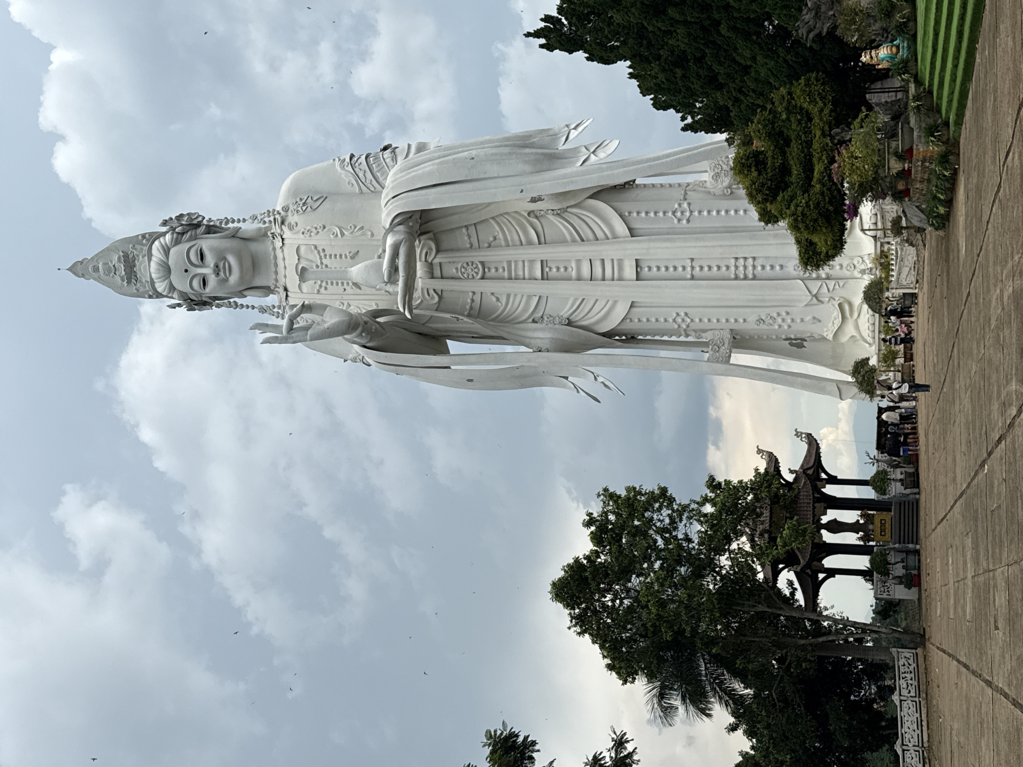 The image shows a massive white statue of Guanyin, a Buddhist deity, standing tall against a cloudy sky. The statue is intricately detailed, with flowing robes and delicate facial features. The statue is located in the city of Nam Ban, Vietnam. In the foreground, there is a paved courtyard with people walking around. To the left of the statue, there is a traditional Vietnamese temple with a multi-tiered roof. The temple is surrounded by trees and lush greenery. The overall scene is one of peace and serenity, with the majestic statue serving as a focal point.