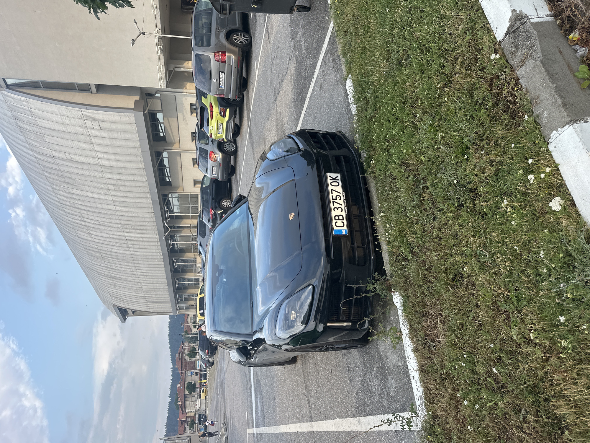 In Samokov, Bulgaria, a black Porsche Panamera is parked in a lot adjacent to a large, curved, silver-roofed building. The car's license plate reads "CB 3757 OK". Other vehicles, including a yellow Hyundai Kona, a grey Volkswagen Touran, and several SUVs, are parked in the background. A few individuals are visible walking in the distance, near some buildings and trees. The sky is partly cloudy. The foreground features a strip of grass with small white flowers and a concrete curb.