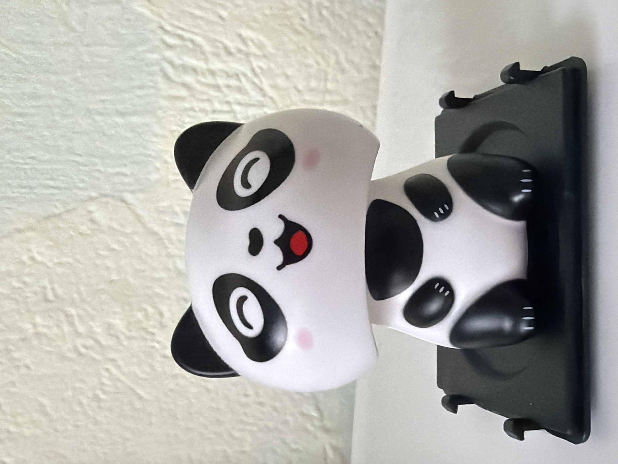 A close-up shot features a cartoonish panda figure seemingly stuck in a flat, black, rectangular surface mounted on a wall. The panda's head is white with black ears, eye patches, a nose, and a smiling mouth. Pink blush marks are visible on its cheeks. The rest of the body is black and white, and the black surface appears to have clips at the bottom. The wall behind the panda is textured and off-white. The location is not discernible from the image.