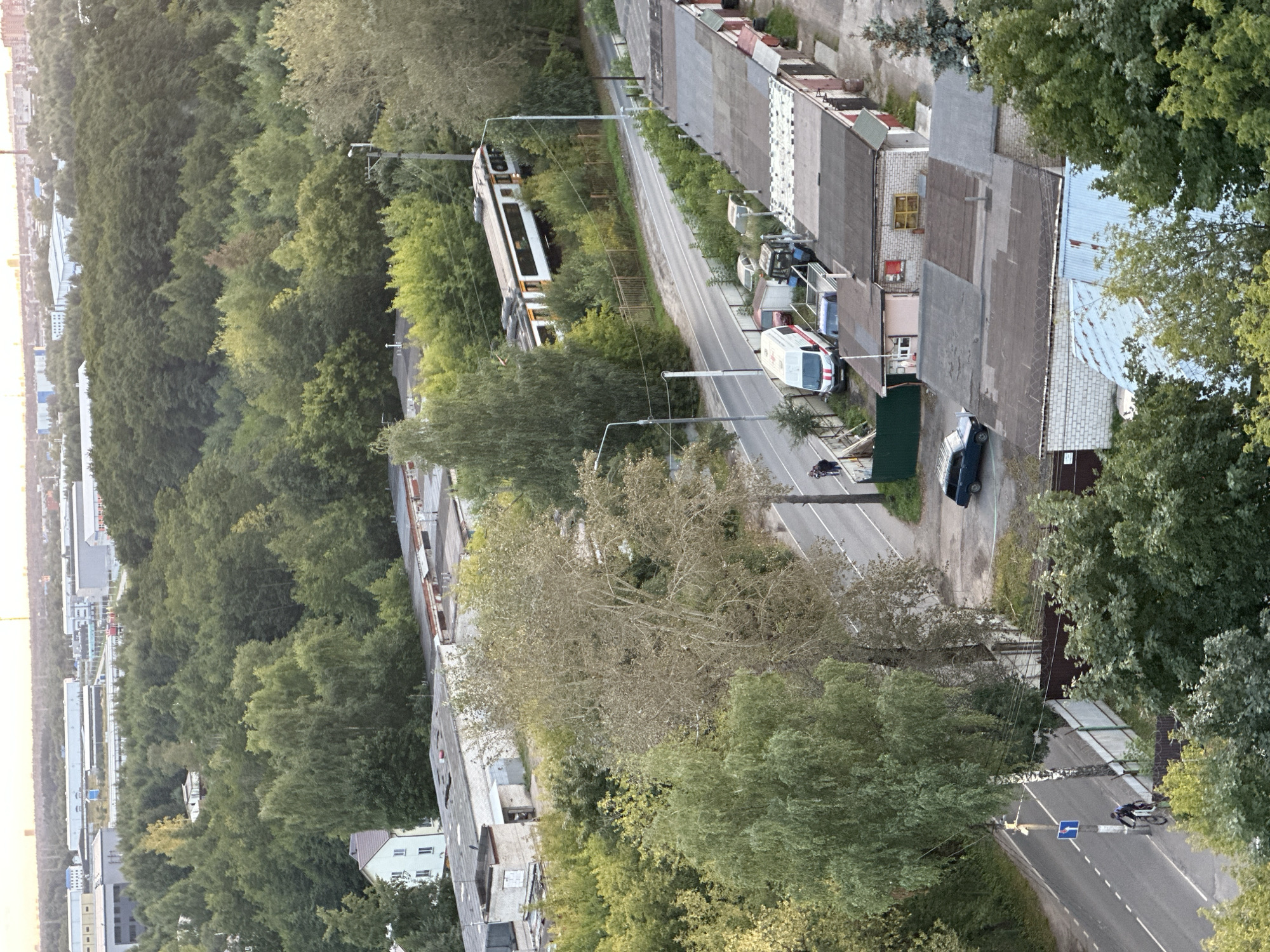 The image depicts an aerial view of a suburban area in Shchyolkovo, Russia, characterized by a dense urban forest bordering a mixture of residential buildings, industrial structures, and garages. The time appears to be late afternoon or early evening, judging by the soft, diffused light.
In the foreground, a row of gray-roofed garages runs horizontally across the image, with some showing signs of wear and tear. Various vehicles are parked or stored in front of these garages, including a white van with red markings, a blue tractor, and several cars. A blue sedan is parked further down the road.
A paved road curves through the scene, with a cyclist in dark clothing riding on the left side of the road, adhering to traffic rules. Further up the road, two pedestrians are visible, walking together. A street lamp stands by the road.
A train, appearing to be a light rail or tram, is moving on a track that runs through a band of trees. The train is white with yellow accents and has its lights on.
The middle ground is dominated by a thick expanse of green trees, indicative of a forested or heavily wooded area. Within the trees, several buildings are partially visible, including a white residential building with a red-tiled roof and a larger, light-colored industrial-style building.
In the background, a more distant urban landscape with various industrial buildings and structures is visible against a hazy sky. The overall impression is one of a transition zone between a natural, wooded environment and a developed industrial and residential area.