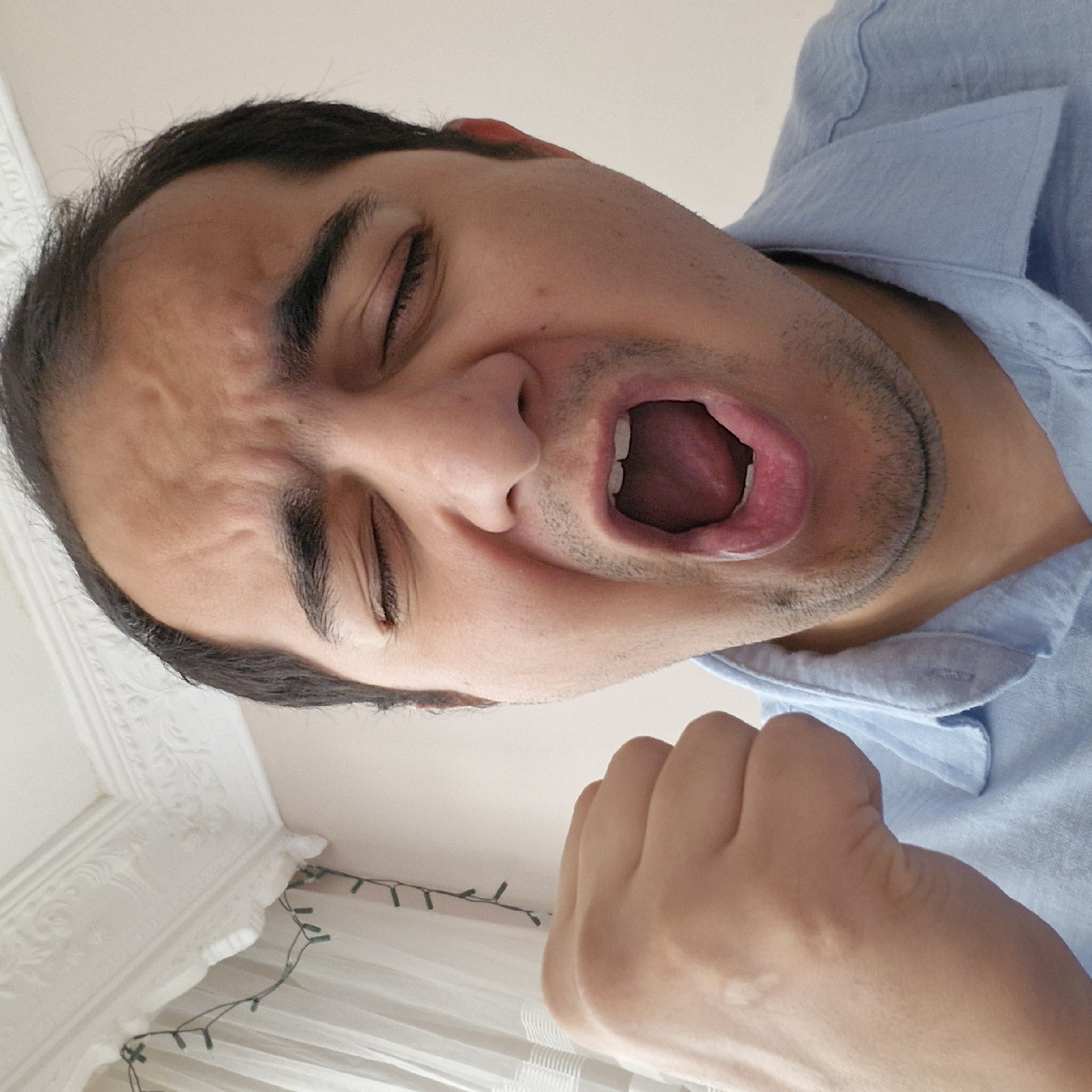 The image is a close-up, selfie-style photograph of a man's face and upper body. The man appears to be yawning or shouting, with his mouth wide open, eyes closed, and a fist raised in the air. He is wearing a light blue collared shirt. The background is blurred but appears to be an indoor setting with white ceiling molding and a curtain. The lighting suggests it is daytime. There are no visible elements that place this image specifically in the city of Küçükçekmece, Turkey.
