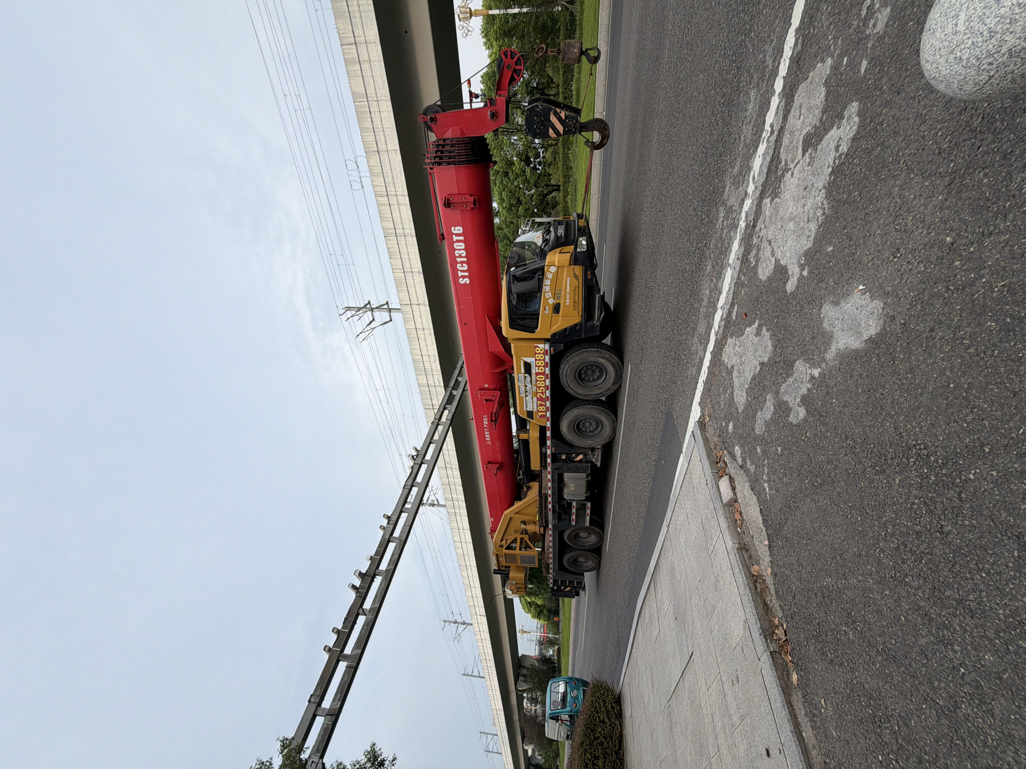 A large red and yellow crane truck is driving on a paved road. The crane is extended. To the left of the crane truck, a small blue vehicle is parked on the side of the road. In the background, an elevated structure with train tracks runs horizontally across the image. The sky is overcast. The scene appears to be in the city of Jiangjin, China, as indicated by the text on the crane truck.
